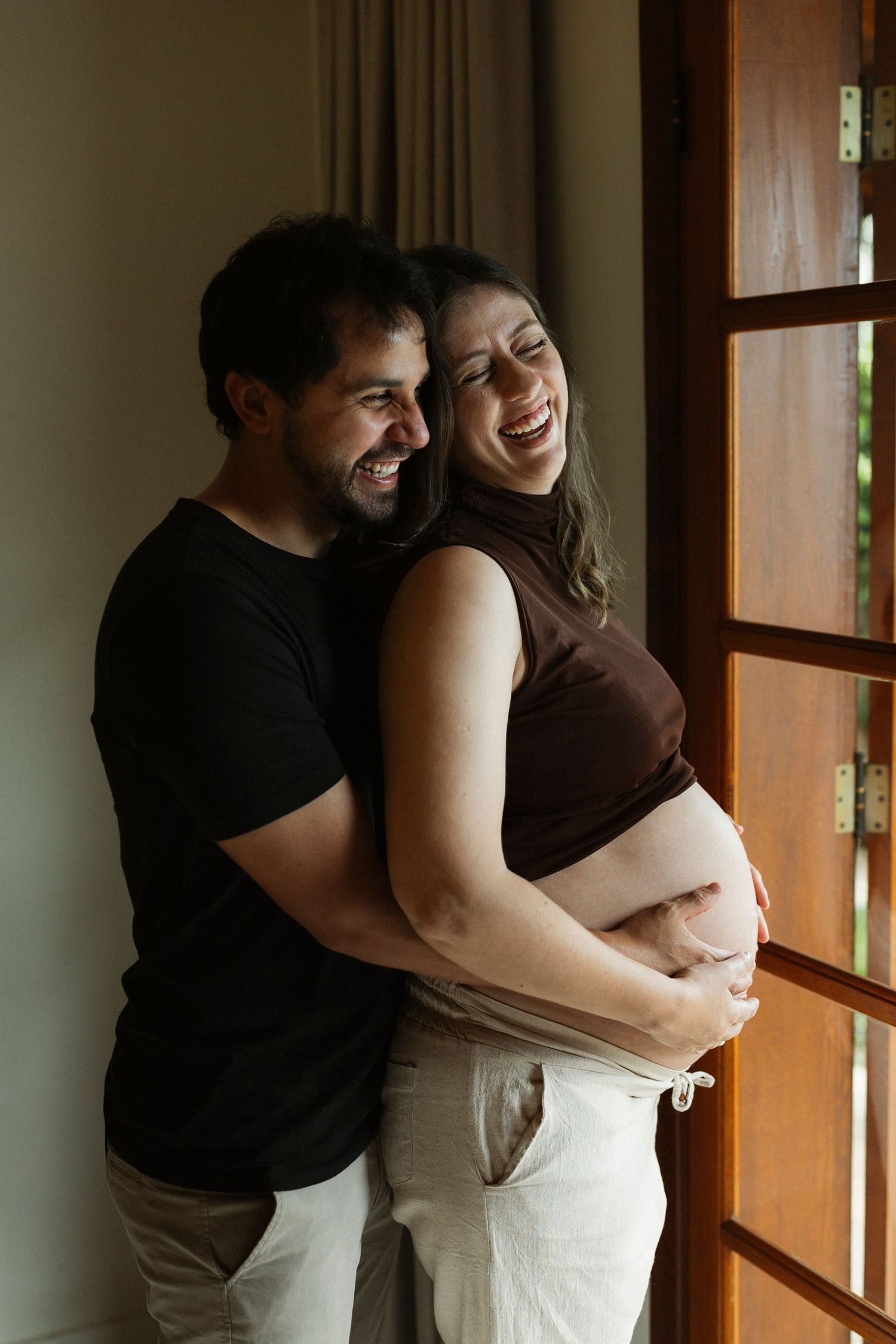 A happy couple laughing together near a window, with the woman visibly pregnant, both dressed casually in black and white.
