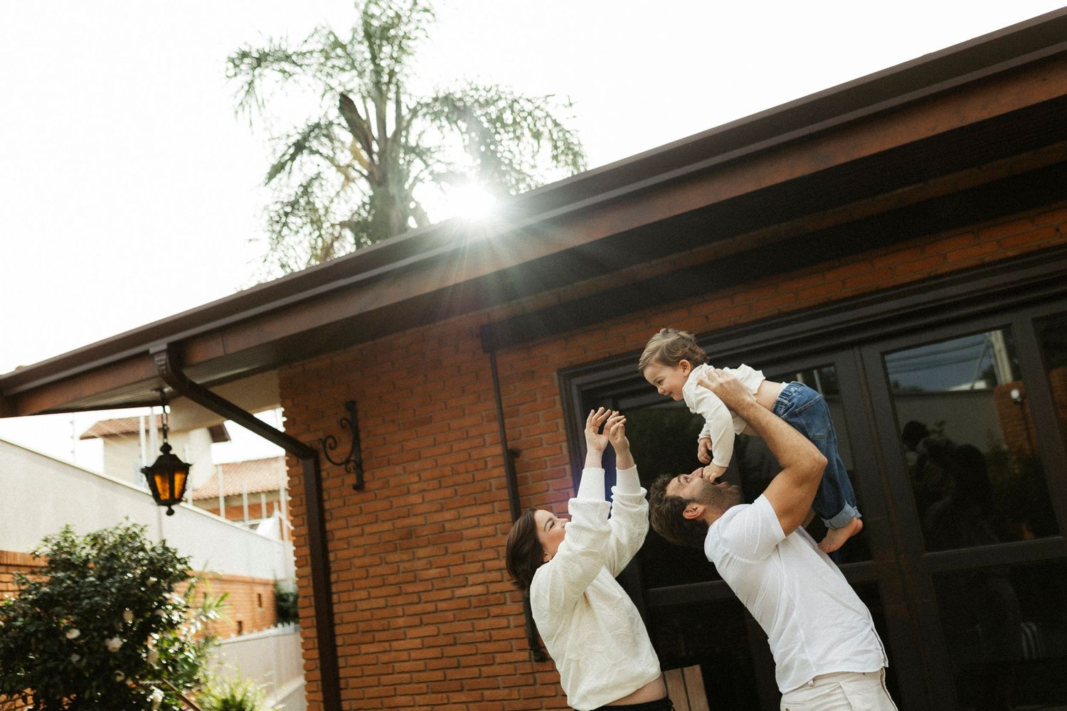 Two adults joyfully lifting a child outdoors near a brick building with sunlight streaming through trees.