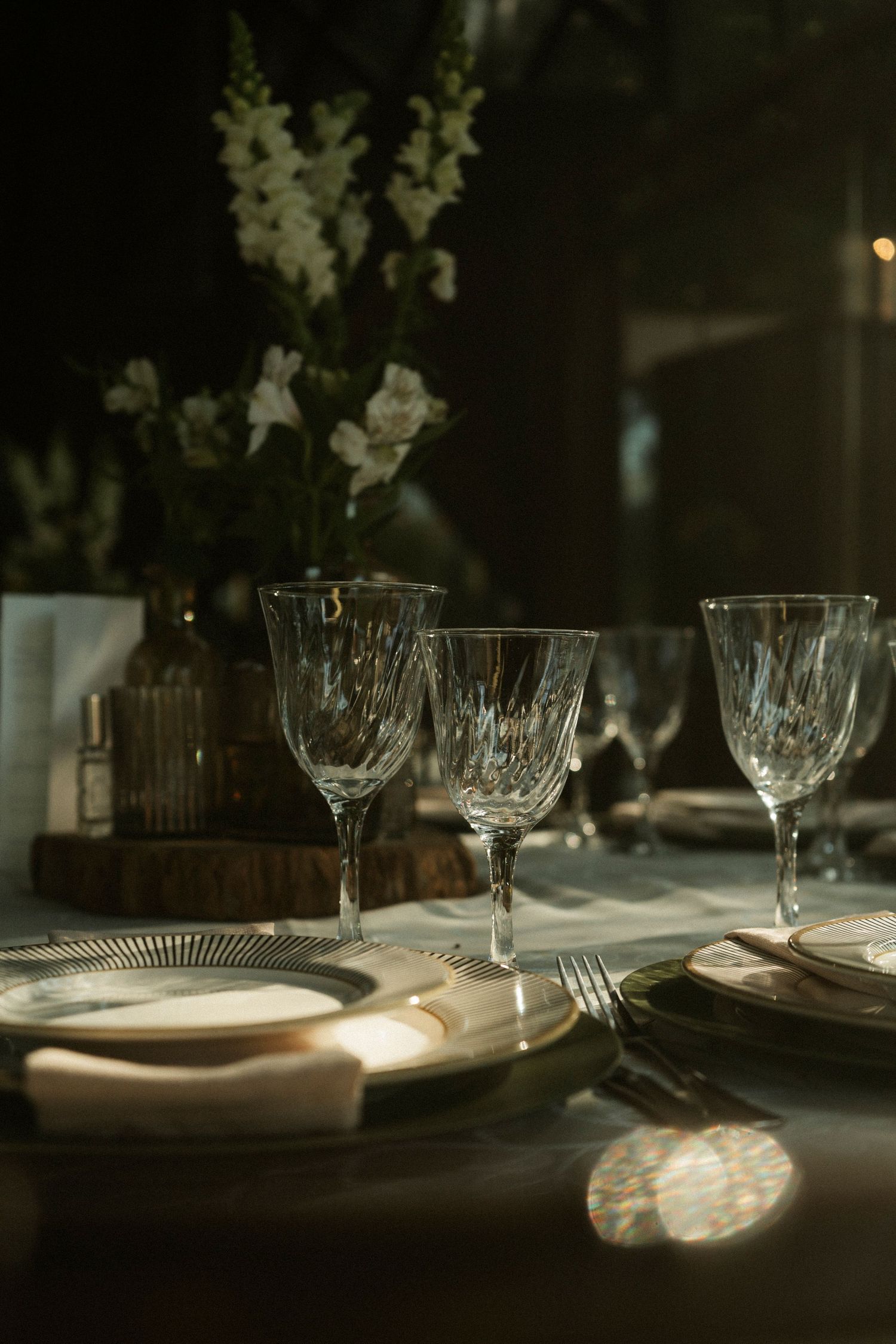 Elegant crystal wine glasses and gold-rimmed plates on a dark moody dinner table with white floral centerpiece.