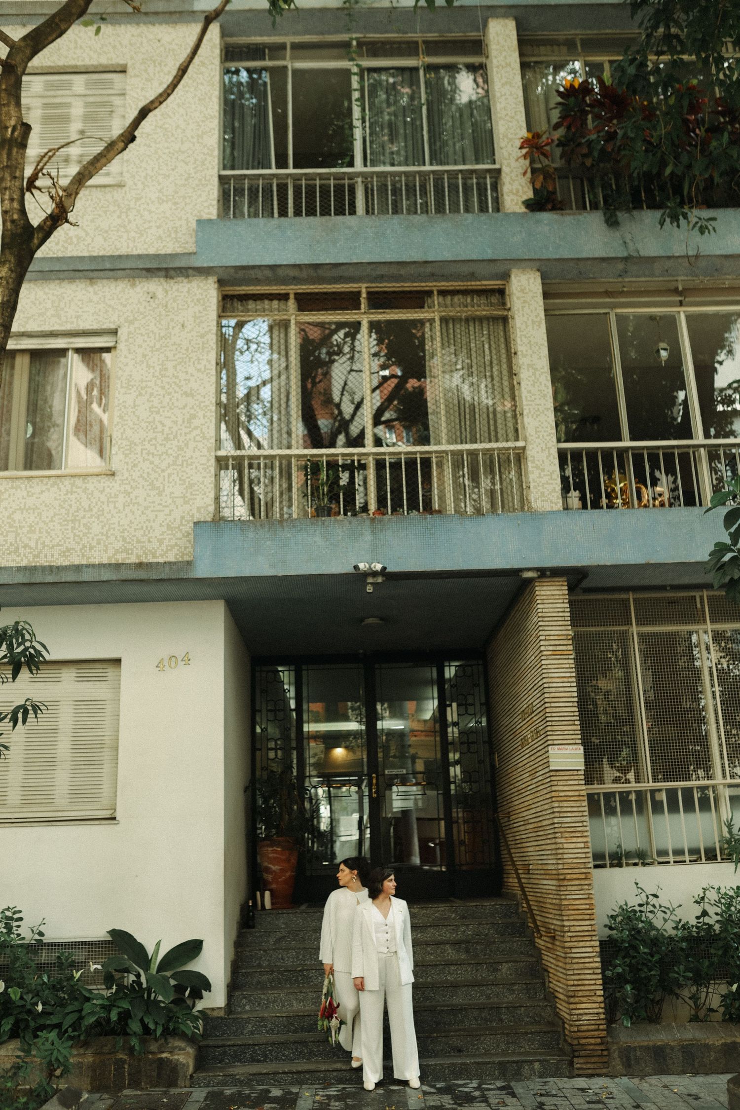 Two people in white clothing stand in front of a modern apartment building entrance.