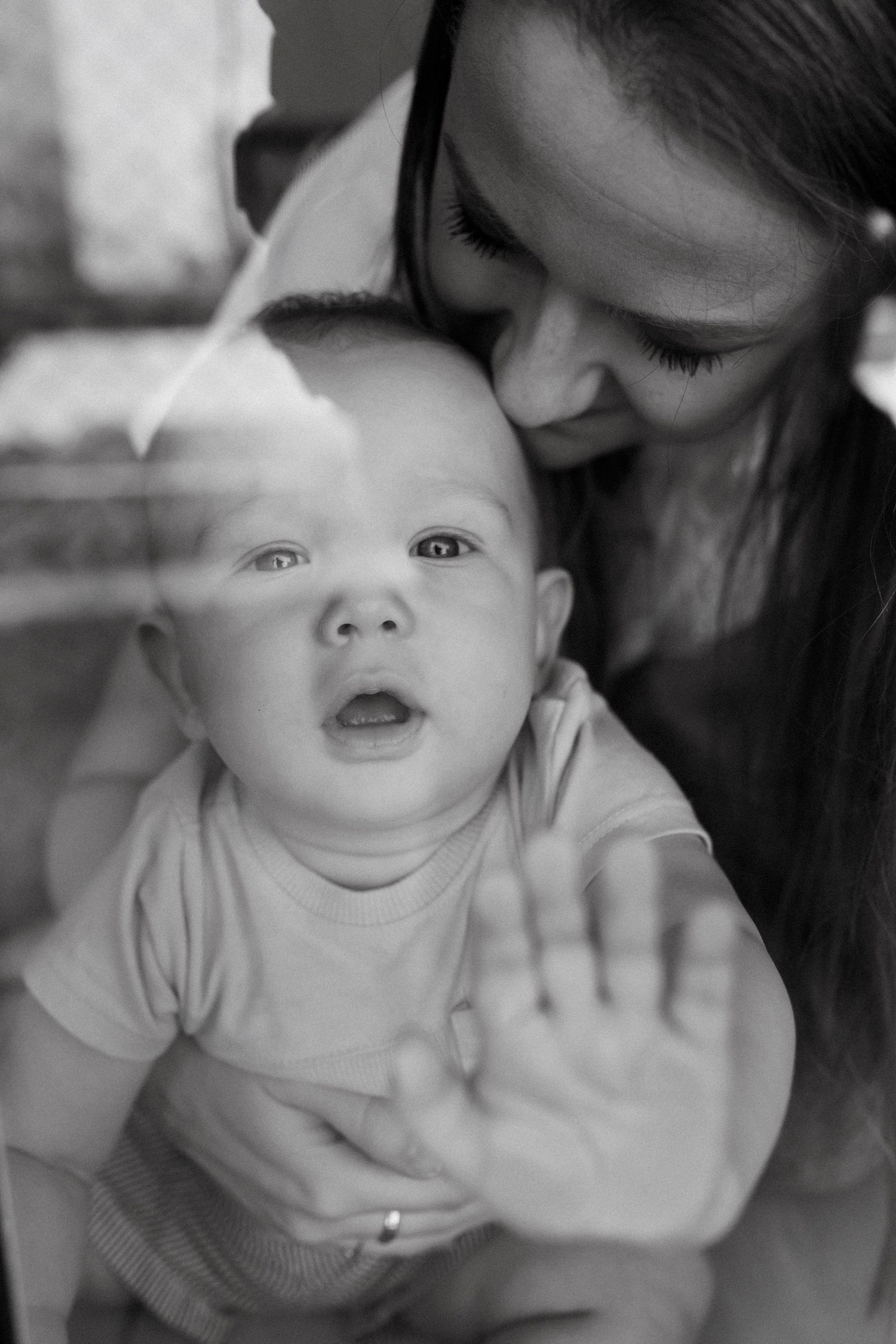 Black and white photo of a mother tenderly holding her curious baby against a window, baby's hand pressed on glass.