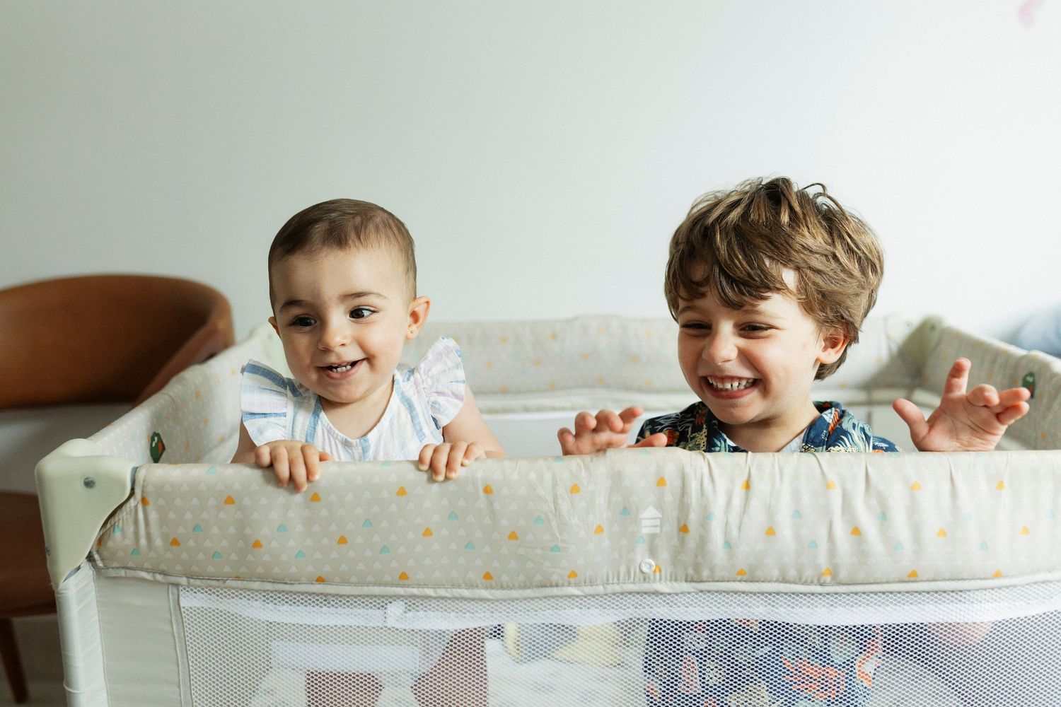 Two young children peek over the edge of a white crib while playing and smiling together.