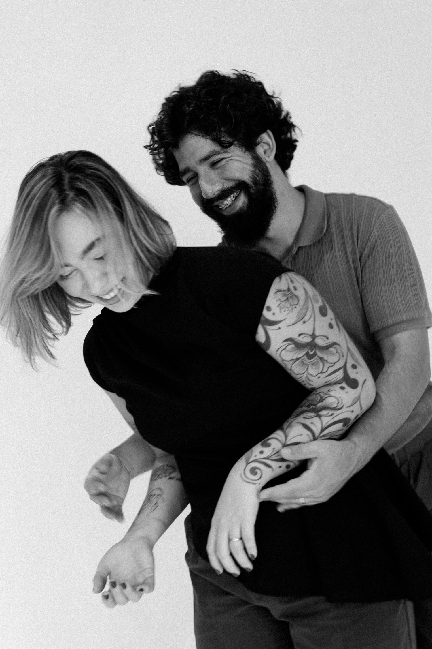 Black and white photo of laughing couple embracing, man with tattoed arm hugging woman in black top.