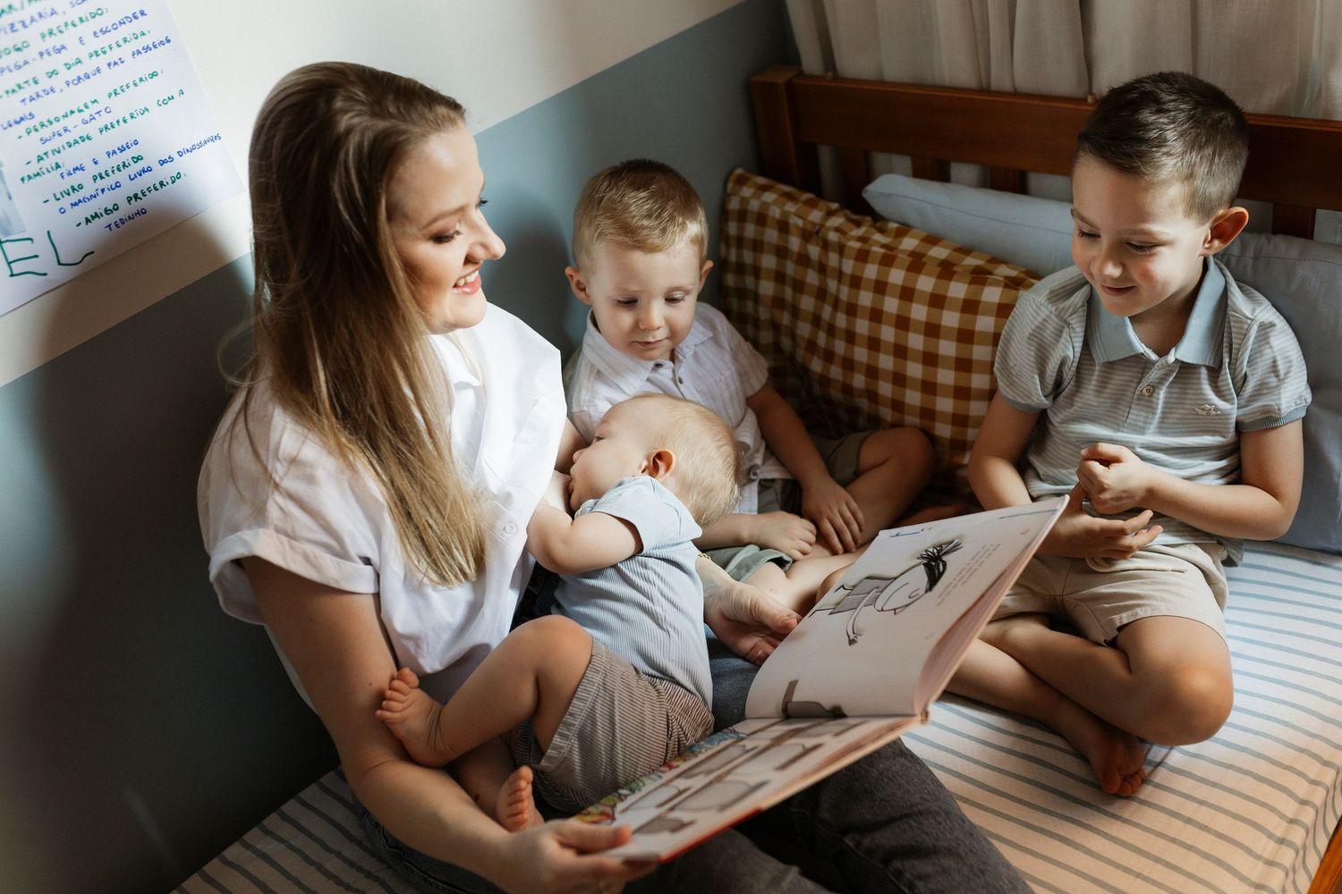 A young woman reads a picture book with two toddler boys on a bed, sharing a warm family storytime moment.