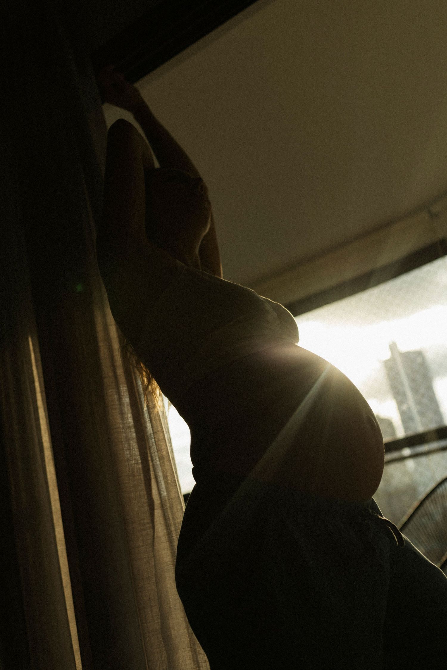 Silhouette of pregnant woman standing by window with golden backlight highlighting her baby bump.