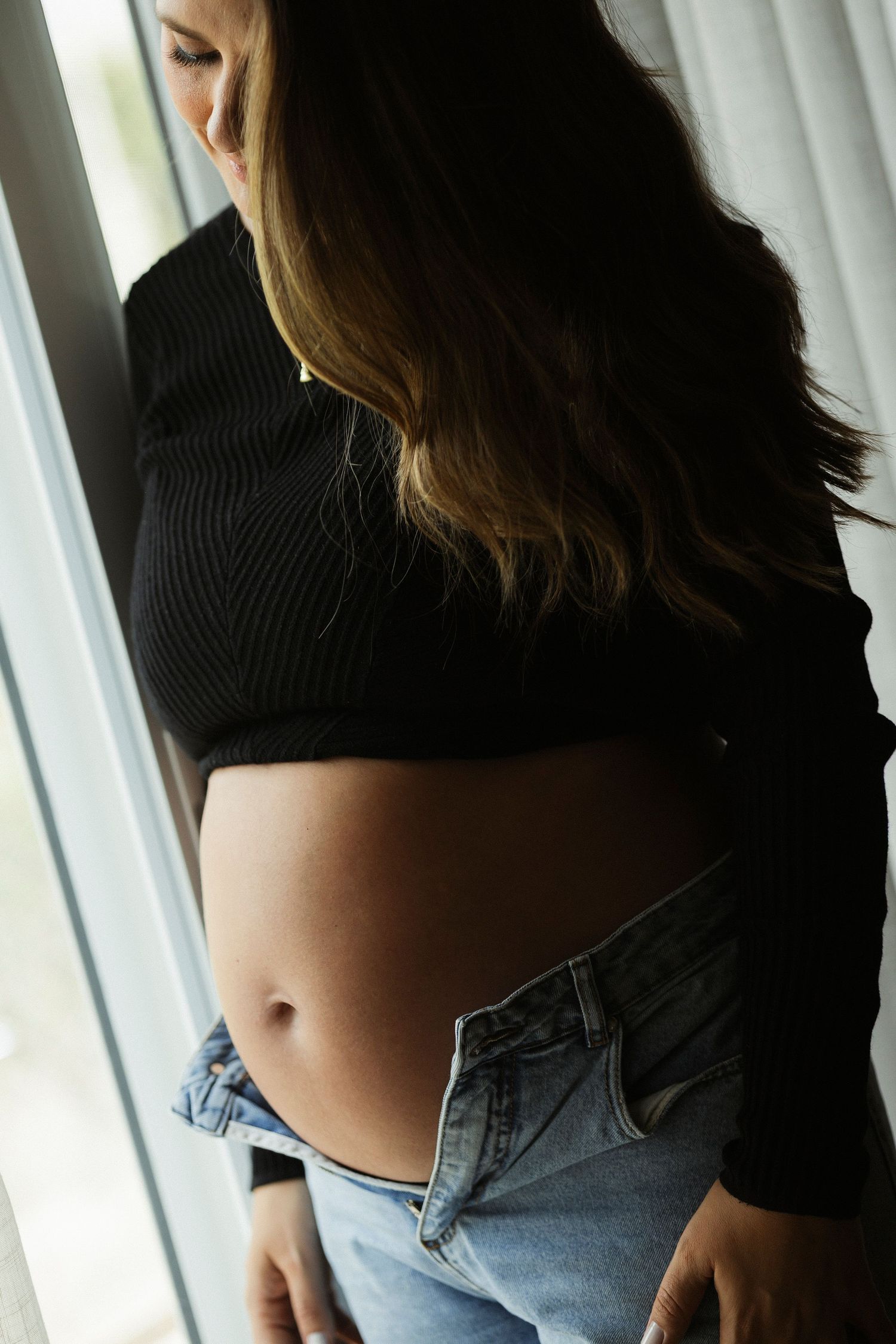 Person wearing a black cropped sweater and jeans poses near a window showing their midriff.