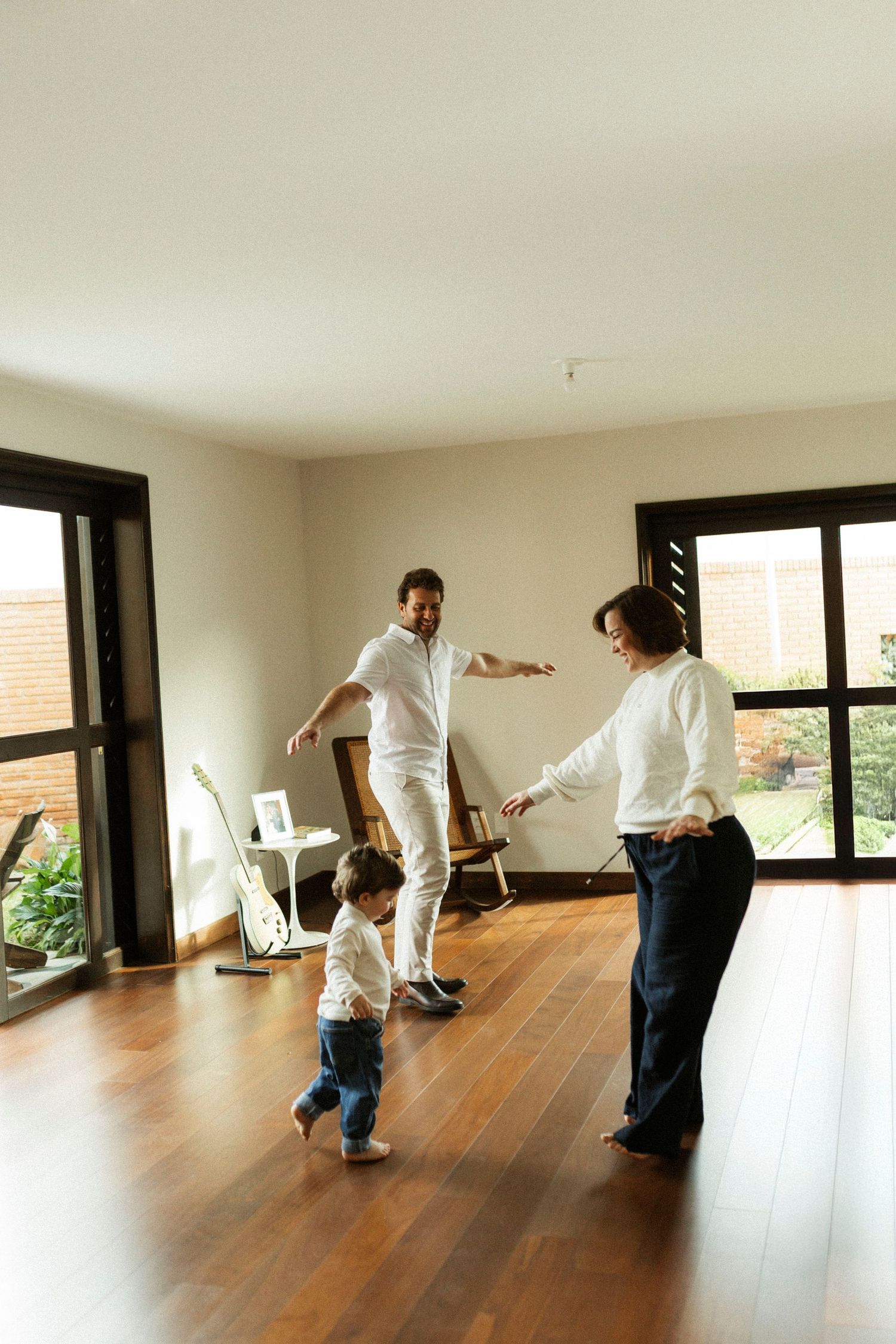 Family with toddler playing together in bright modern living room with hardwood floors and large windows.