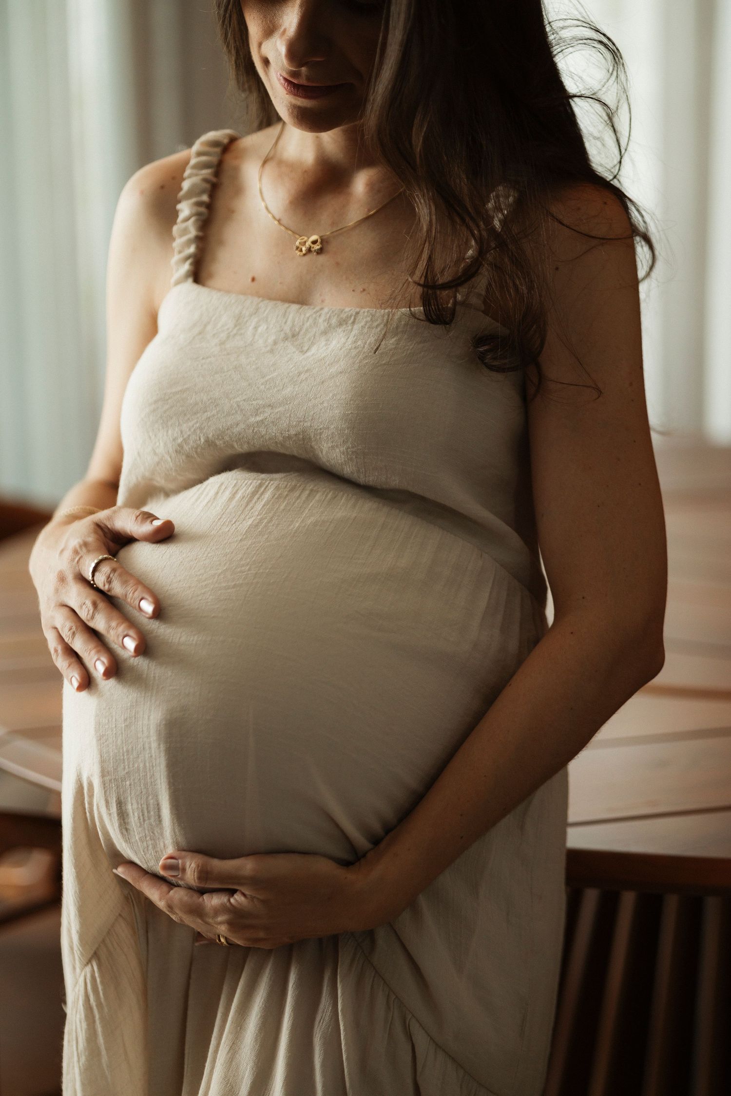 Pregnant woman in white dress gently holding her belly in soft window light.