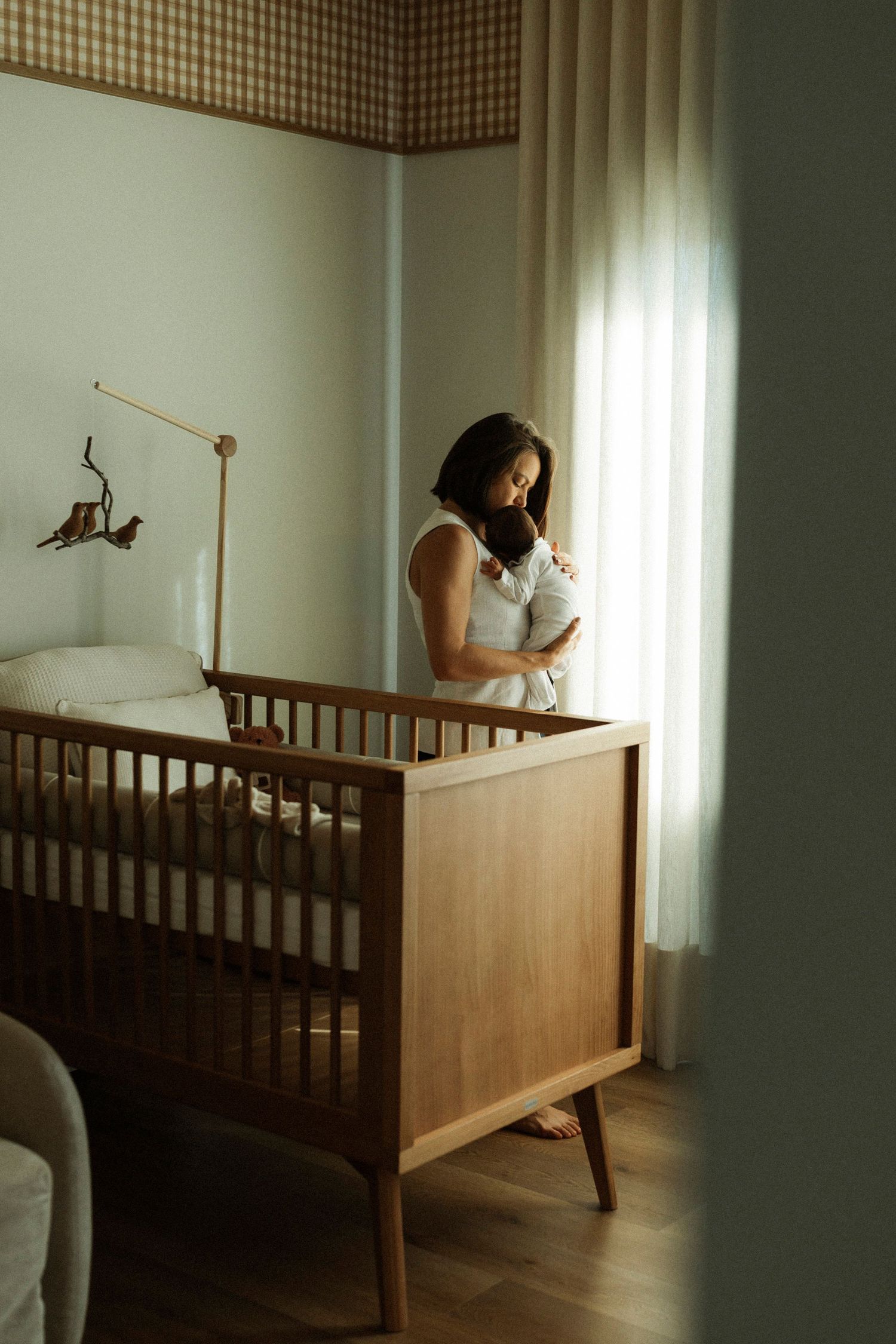 Mother tenderly holds newborn baby beside wooden crib in softly lit nursery with bird mobile.