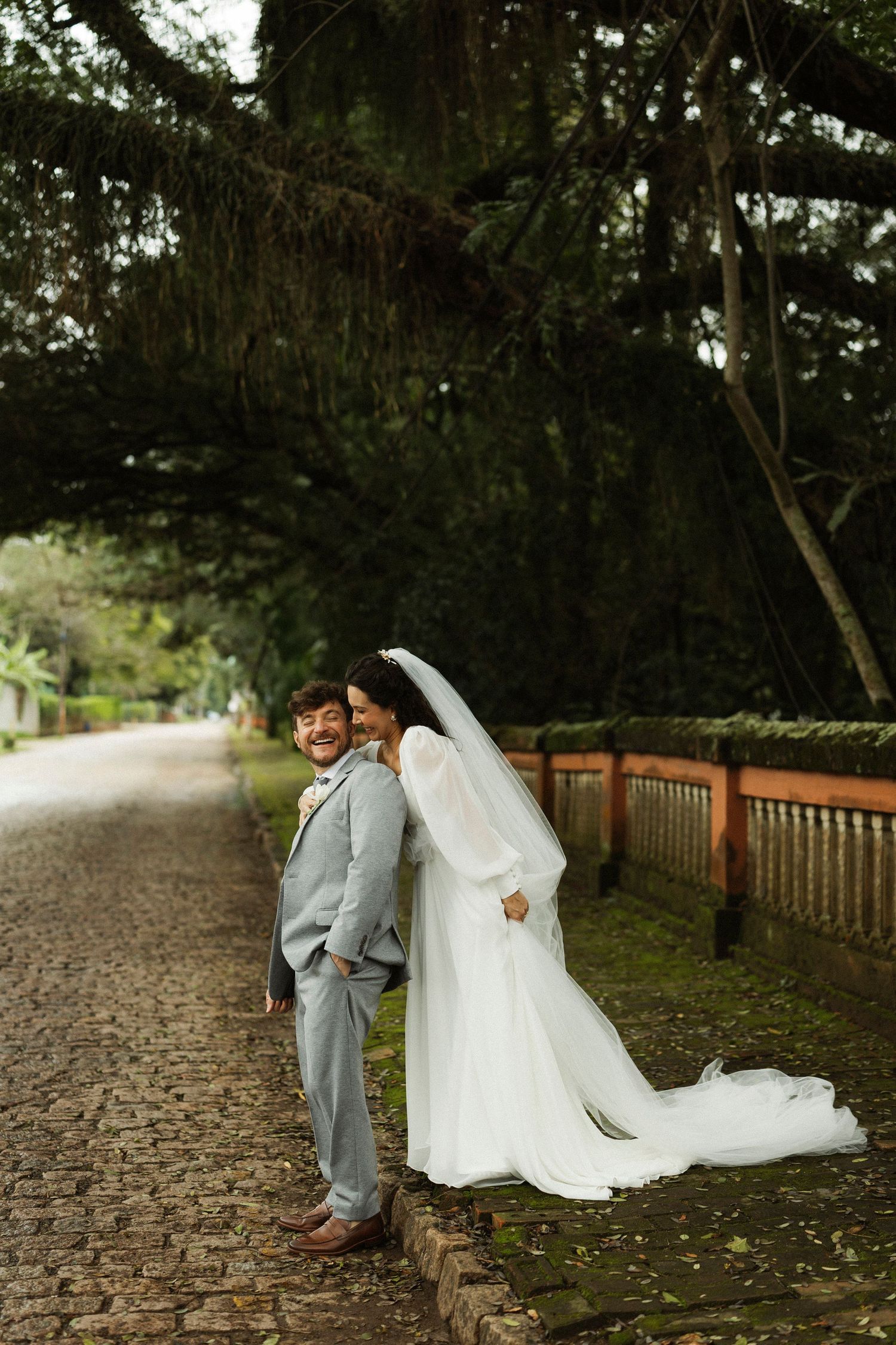 Bride in white gown and veil leans on smiling groom in gray suit on cobblestone path surrounded by lush trees.