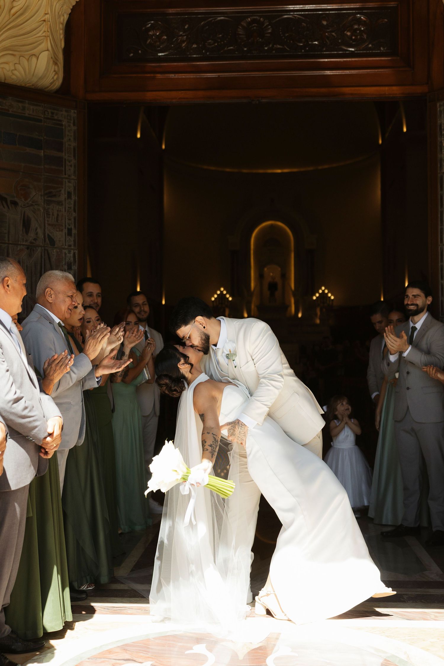 Groom dips bride for a kiss at wedding ceremony exit, guests in green bridesmaid dresses applaud in elegant venue.