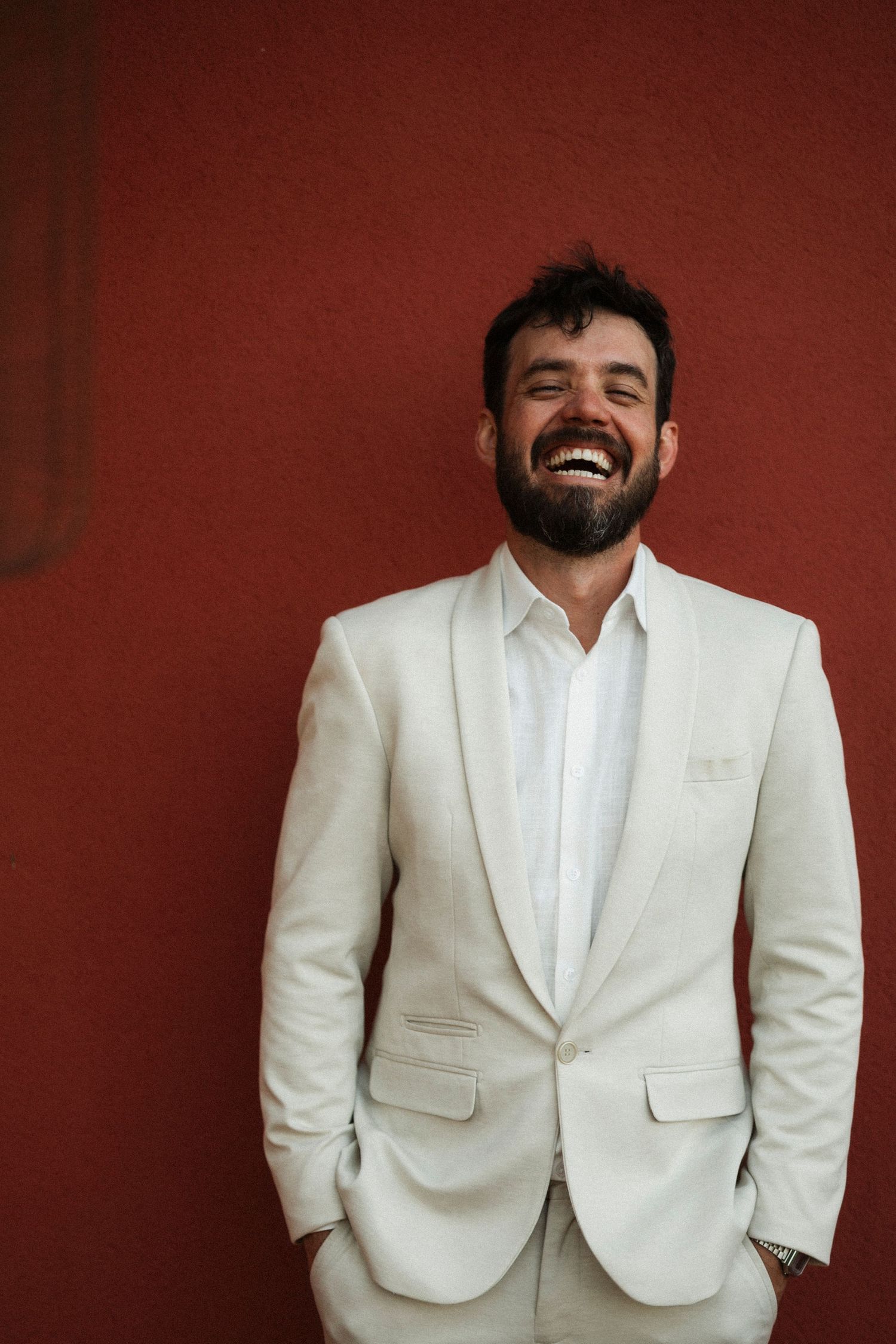 Groom in white suit jacket smiles against burgundy wall during wedding portraits.