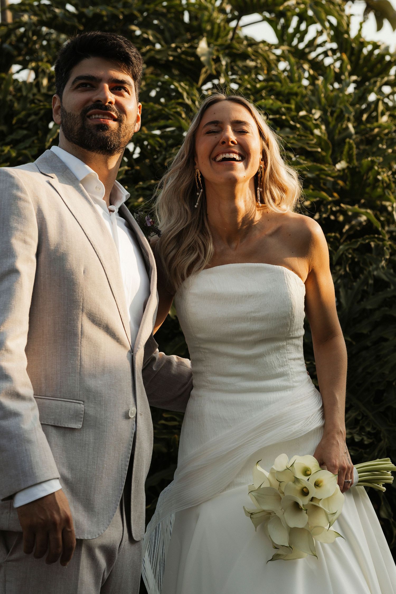 A joyful wedding portrait captured in warm sunlight with tropical foliage in the background.