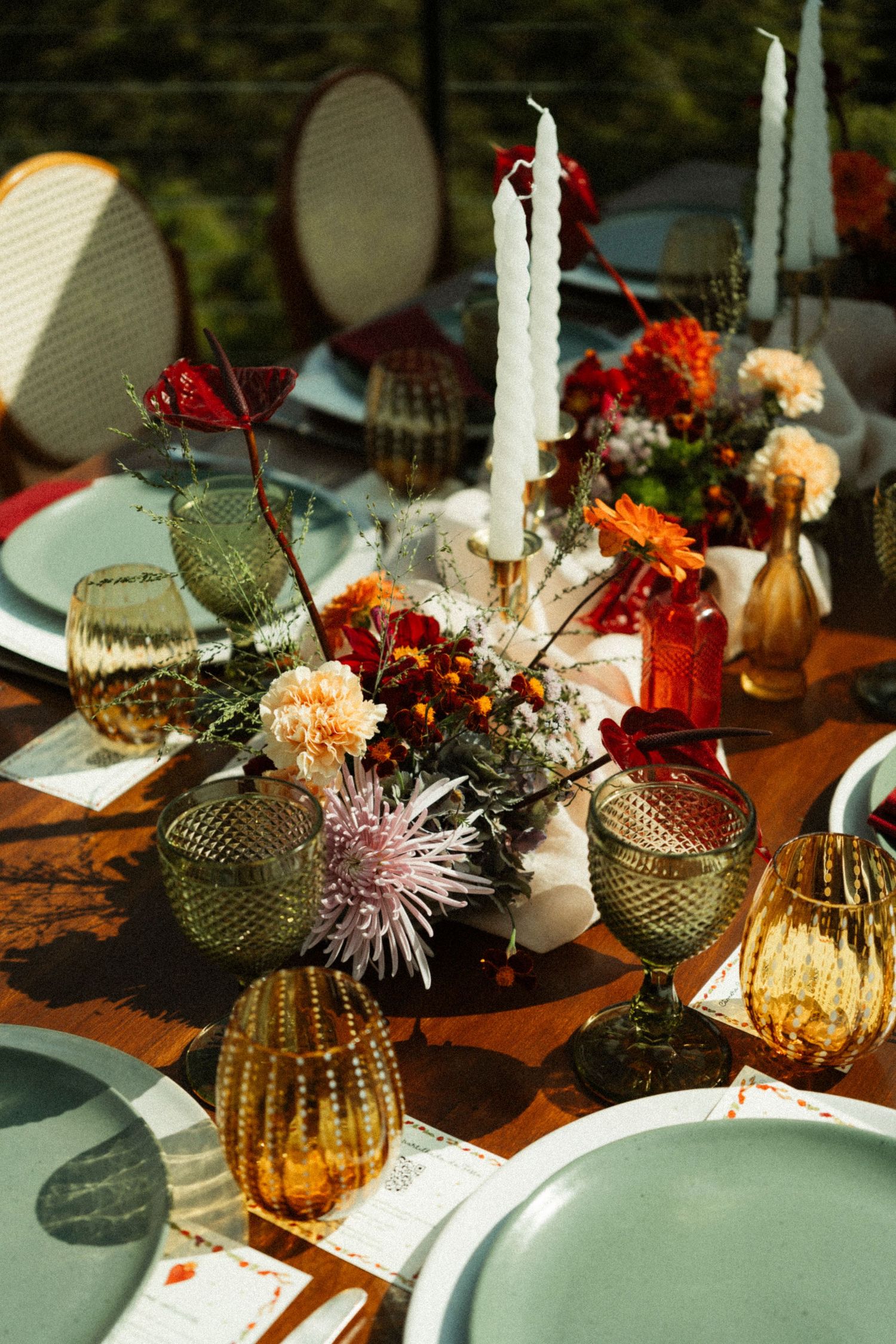 Elegant outdoor tablescape with colorful glassware, white candles, and autumn floral centerpieces on a wooden table.