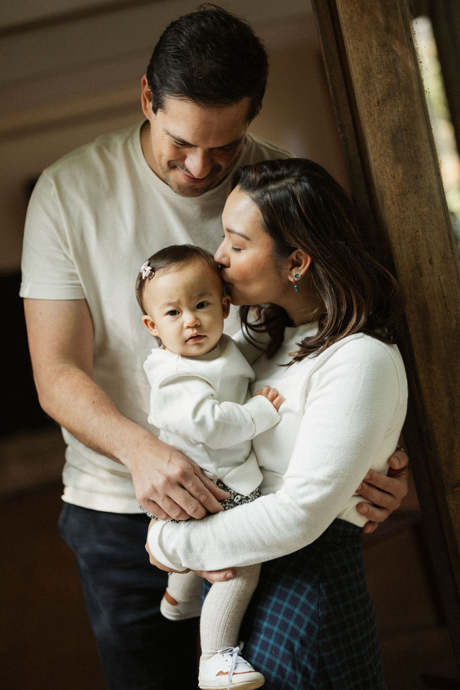 A young family with a mother and father lovingly holding their baby girl, all dressed in white, in a warm indoor setting.