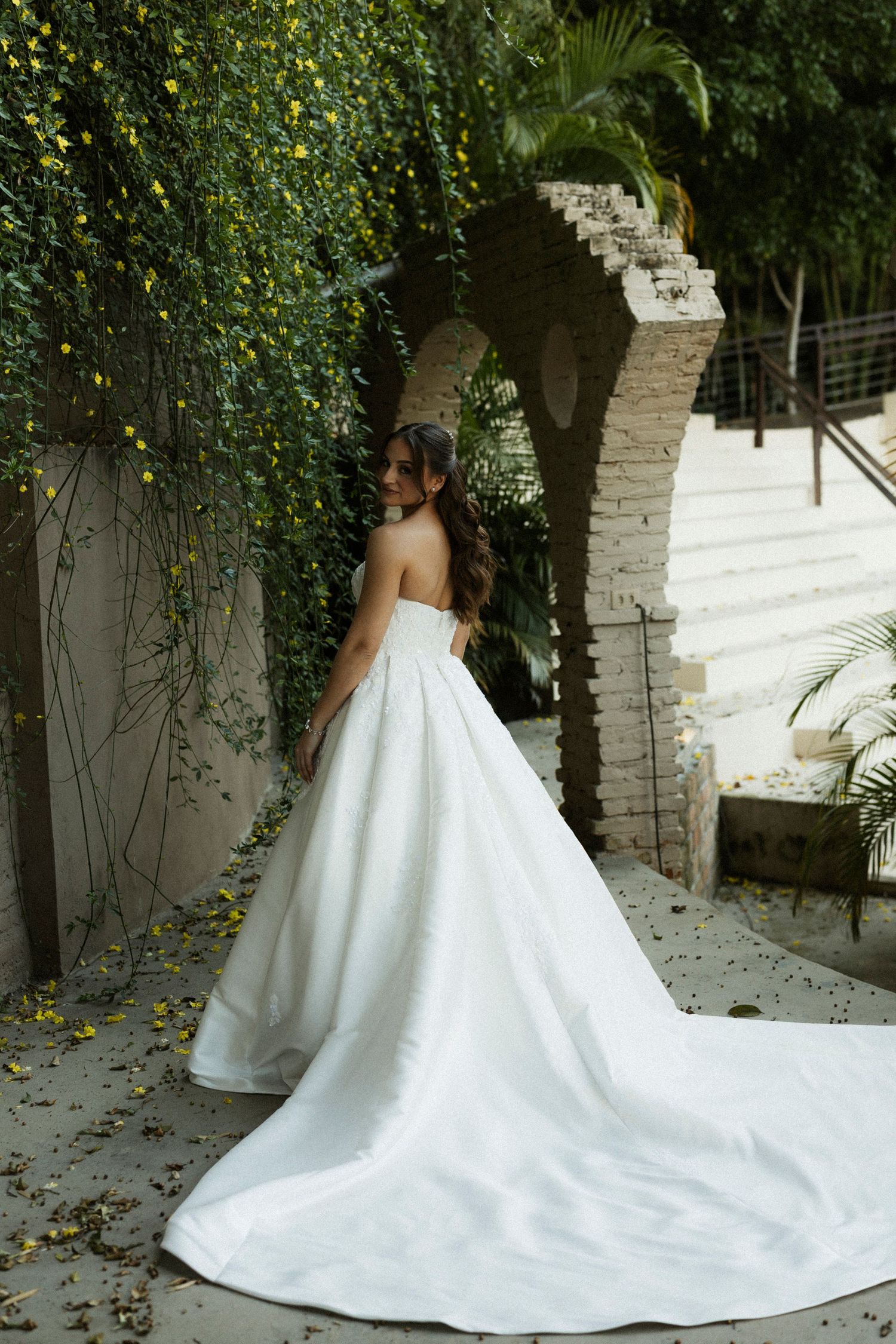 Bride in strapless white ballgown with long train poses near stone arch covered in lush green vines.