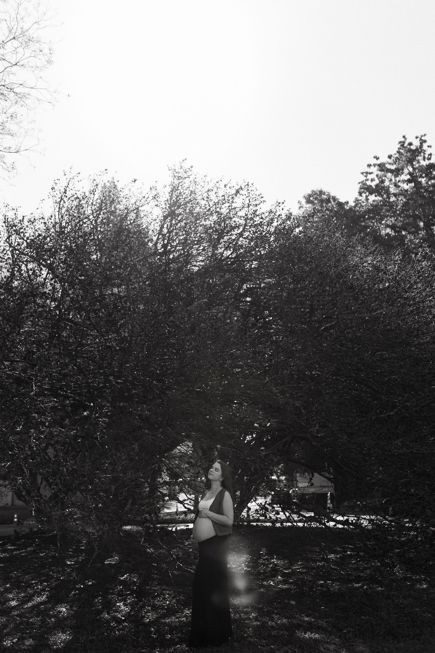 Black and white photo of a pregnant woman standing among trees, looking upward in a serene outdoor setting.