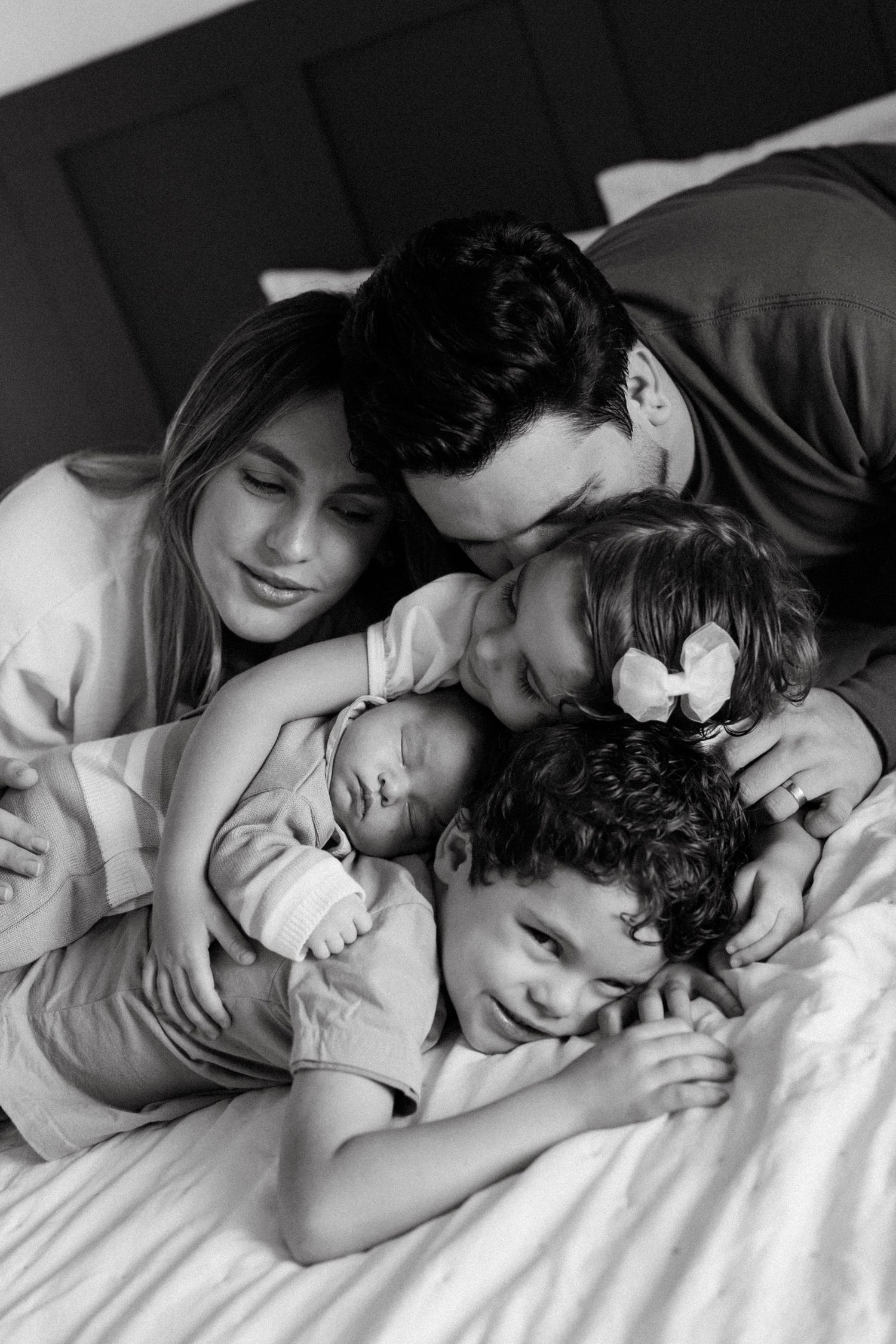 Black and white family photo of parents with three children, including a newborn, cuddling together on a bed.