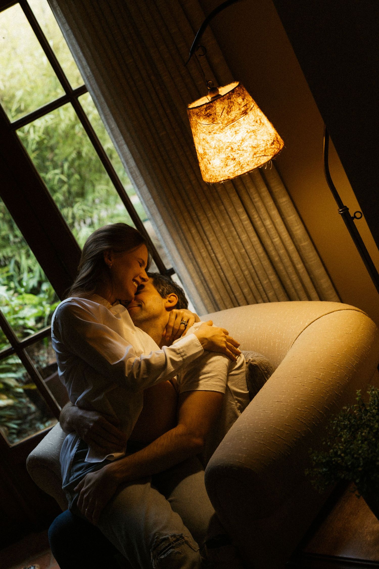 Couple sitting together on couch under warm lamp light in cozy living room with window view.
