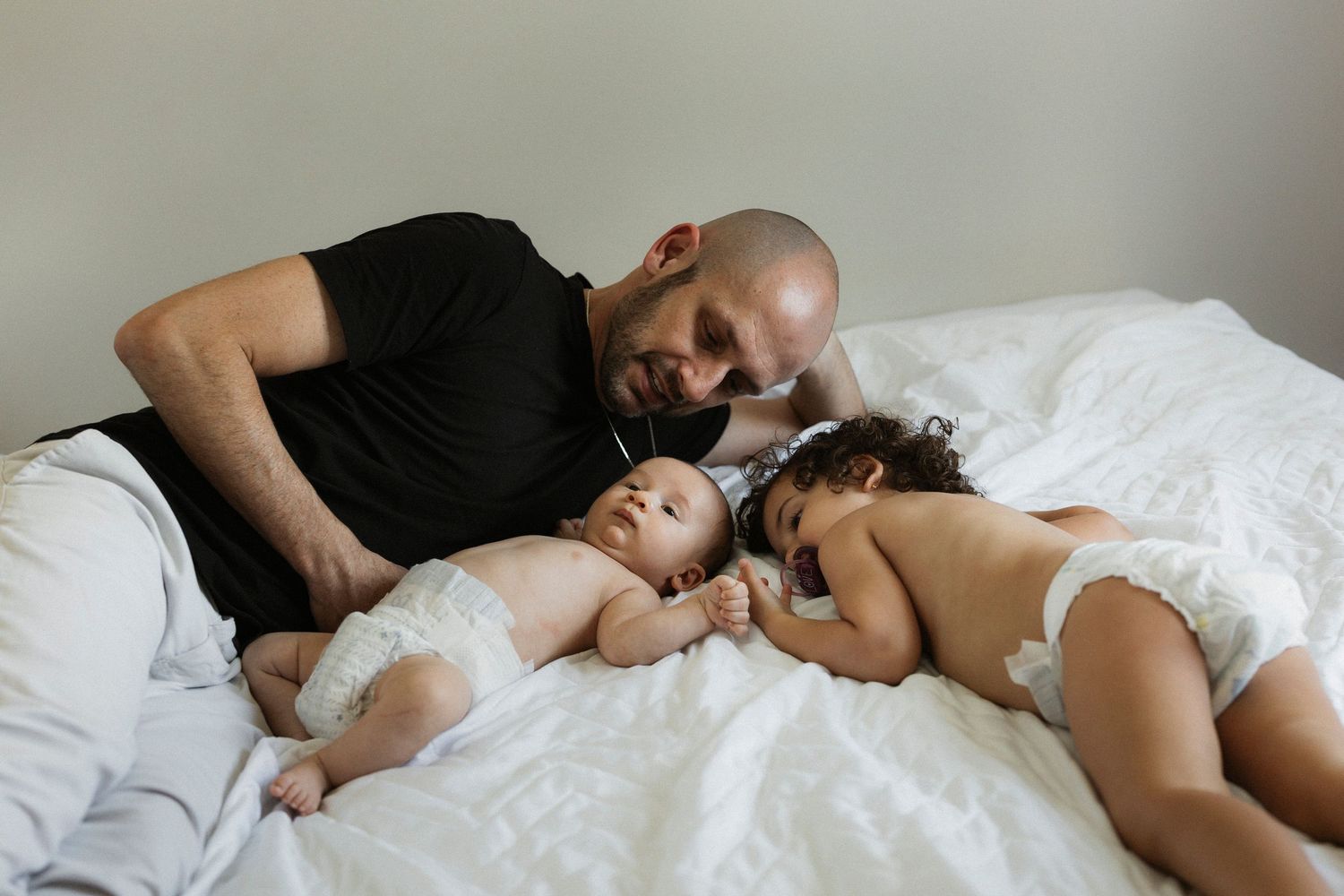 Father lying on white bed with two babies in diapers, sharing a tender family moment together.