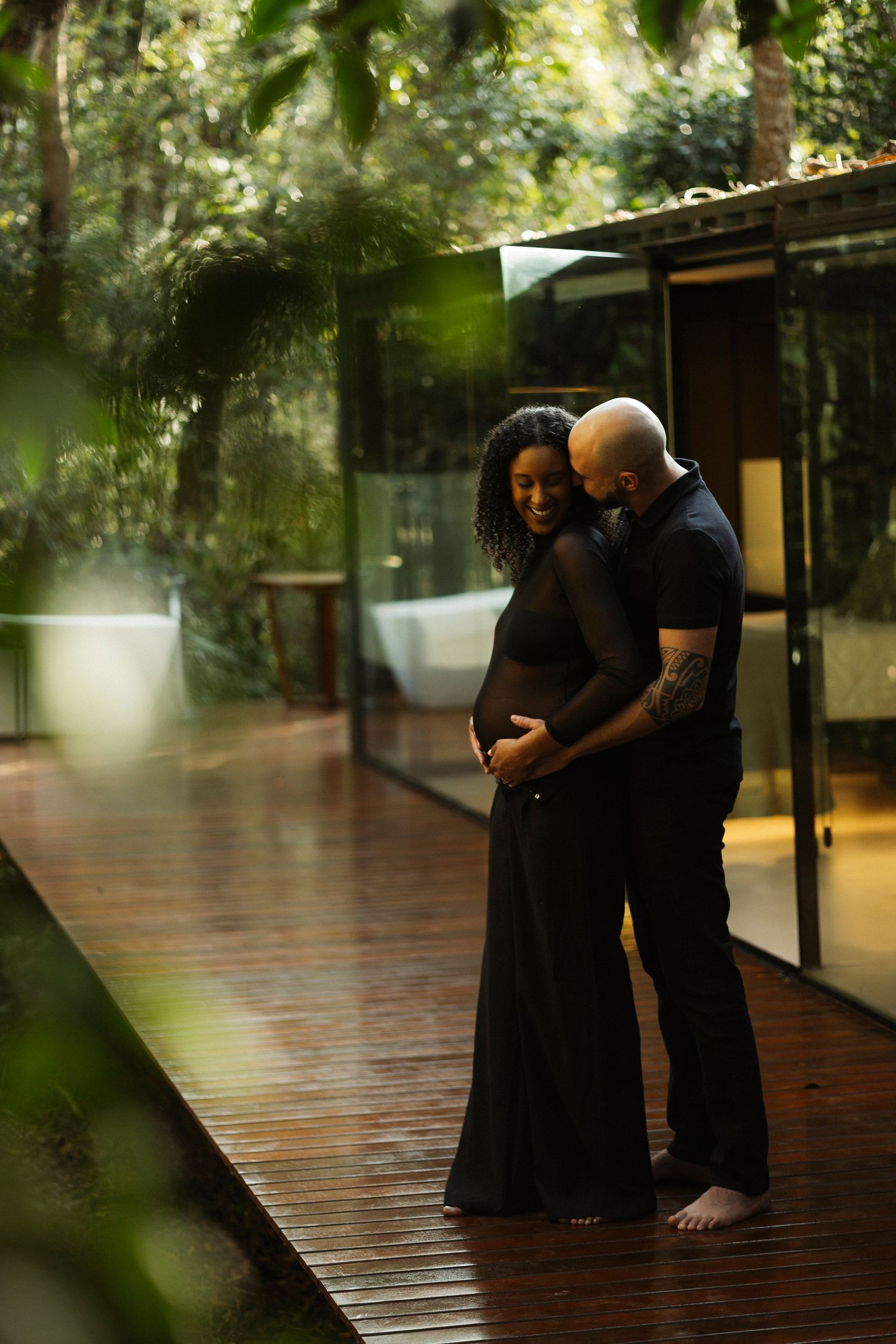 Couple embracing on wooden deck surrounded by lush greenery and natural lighting.
