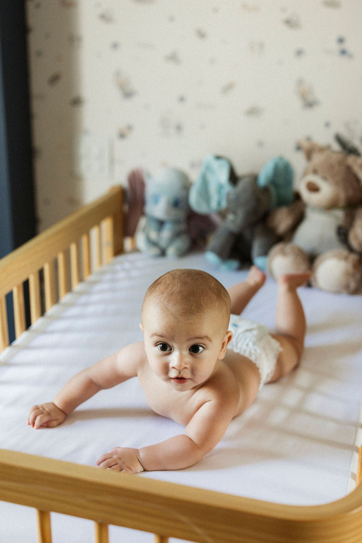 Cute baby in diaper doing tummy time in wooden crib surrounded by stuffed animal toys.