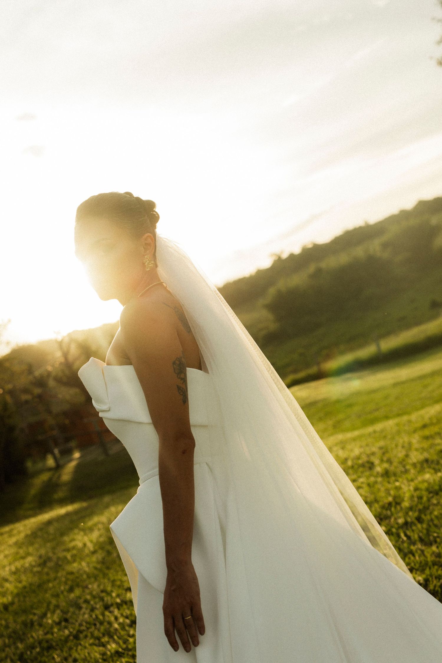 Bride in white wedding dress and veil standing on green hillside at golden hour sunset with natural landscape background.