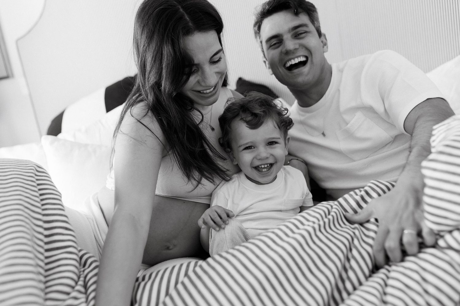 Black and white photo of happy family with toddler laughing together on bed with striped bedding.