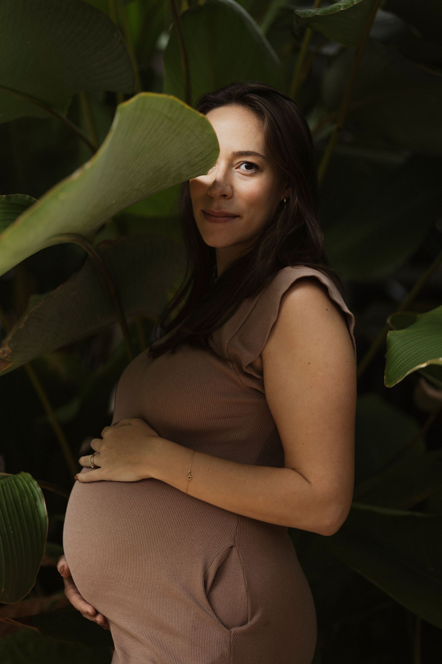 Expectant mother in brown dress poses among large tropical leaves in natural lighting.