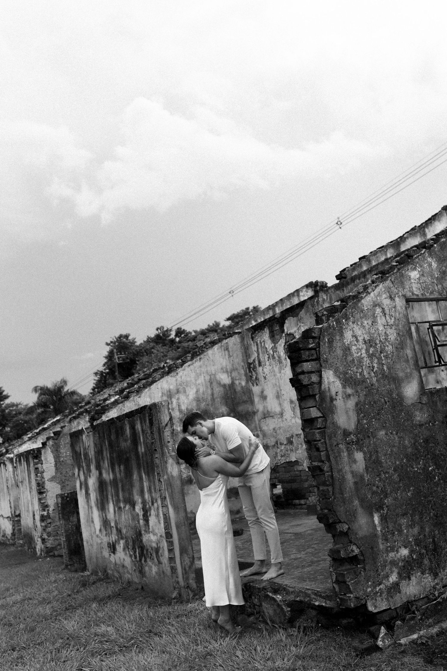 Couple in white clothing posing near weathered concrete buildings in black and white photography.