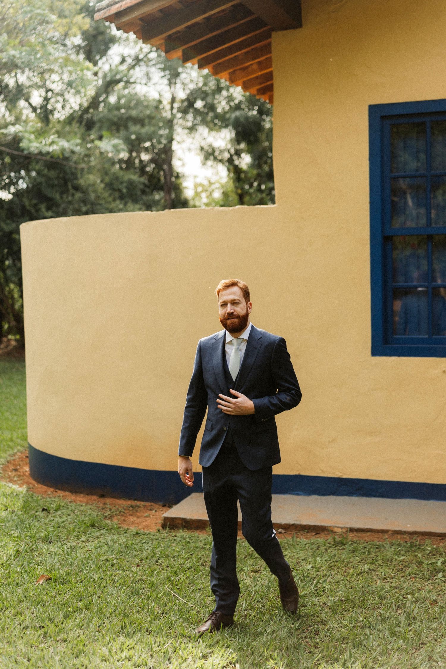 Man in navy suit standing on grass in front of yellow building with blue door and wooden beams.