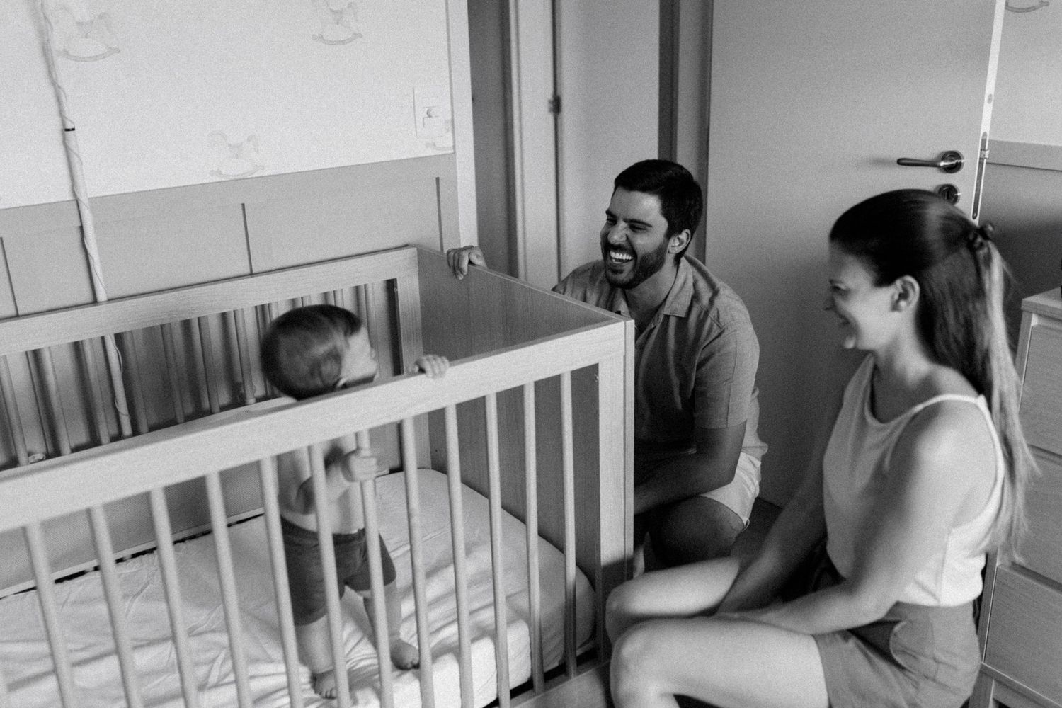A black and white photo of a family sharing a joyful moment together by a crib in a hospital room.