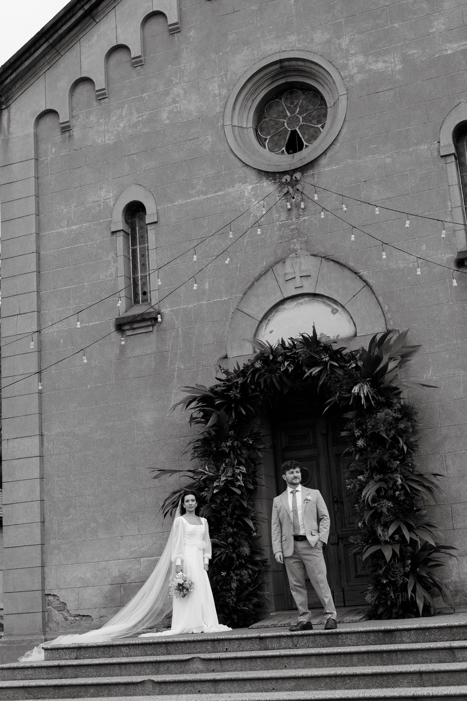 Black and white photo of bride and groom standing on church steps surrounded by lush tropical greenery.