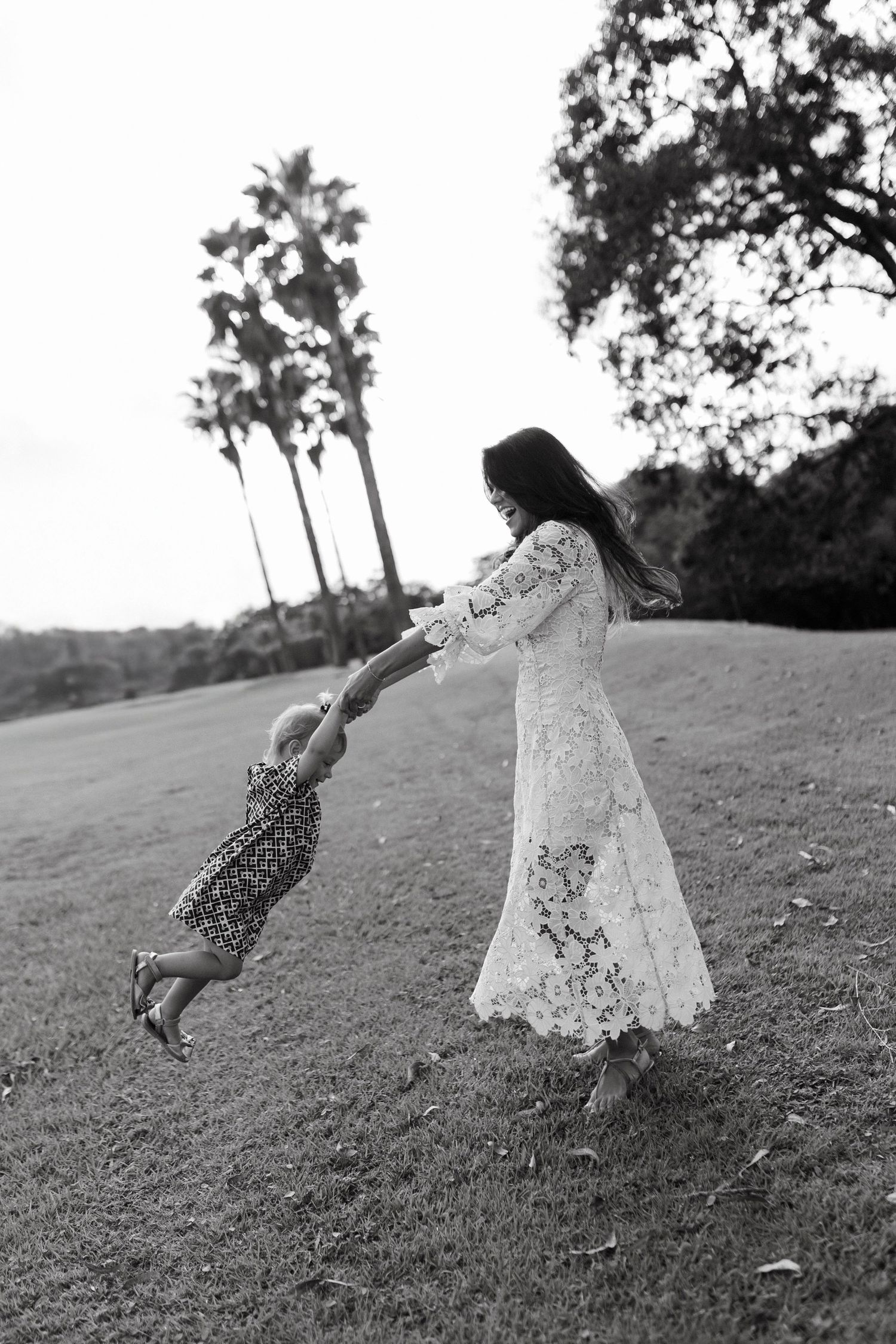 Black and white photo of a mother in lace dress swinging her toddler on a grassy lawn with palm trees.