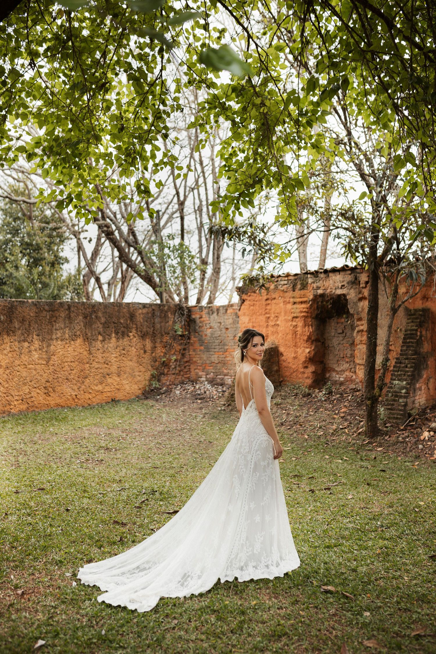 Bride in flowing lace gown poses in garden setting with brick wall and trees.