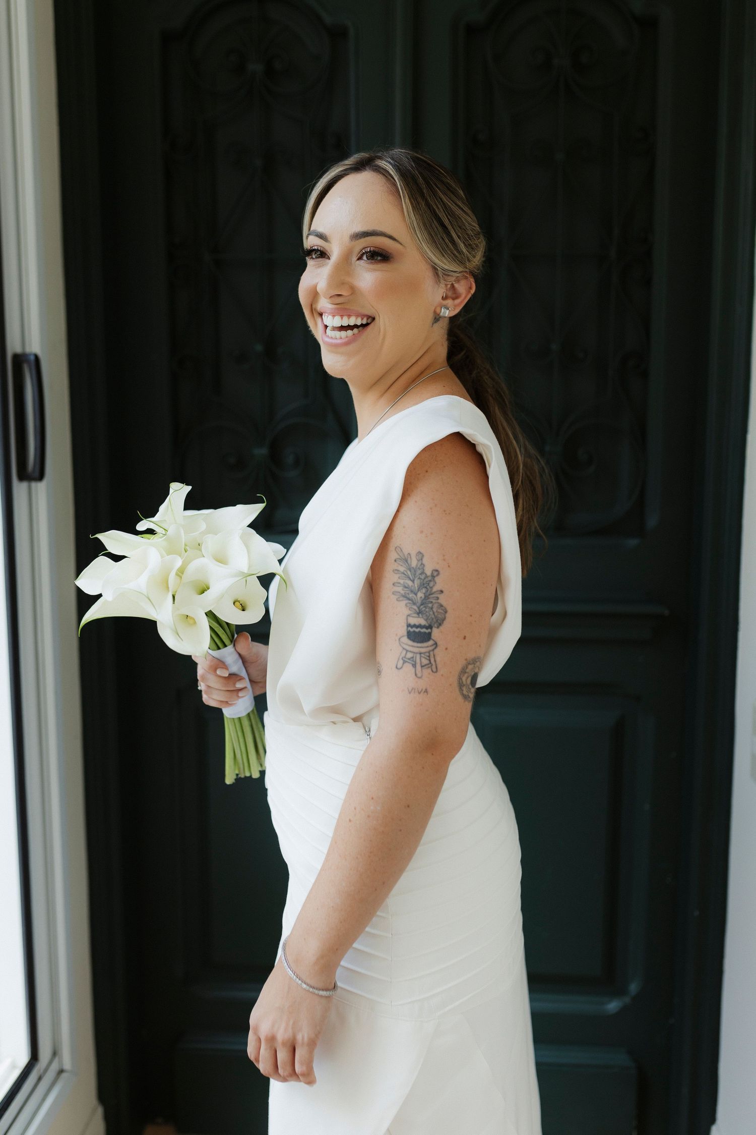 Joyful bride in modern white dress poses with calla lily bouquet by dark doors.