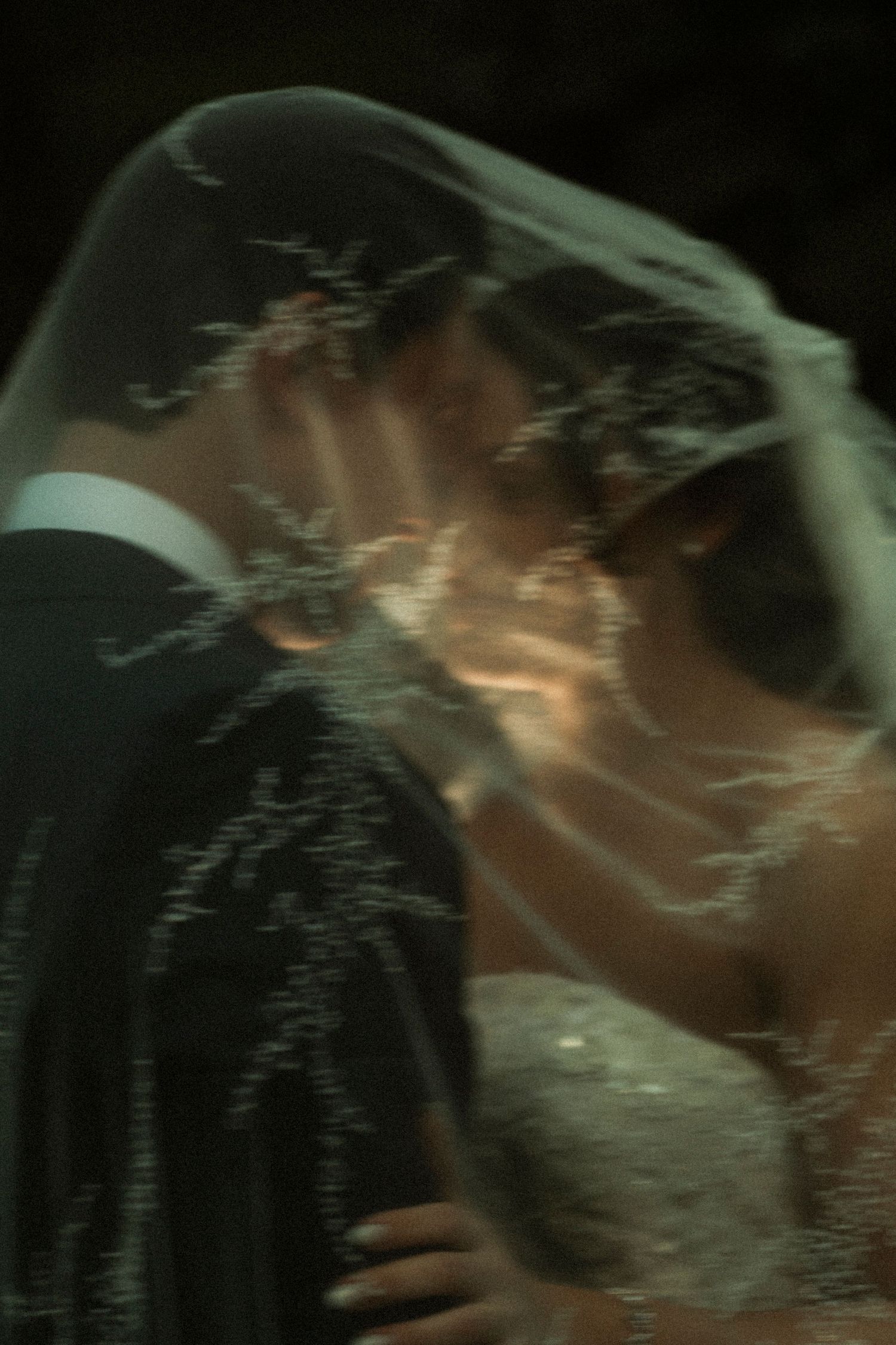 A bride and groom share a romantic kiss beneath a delicate lace wedding veil in a softly lit portrait.