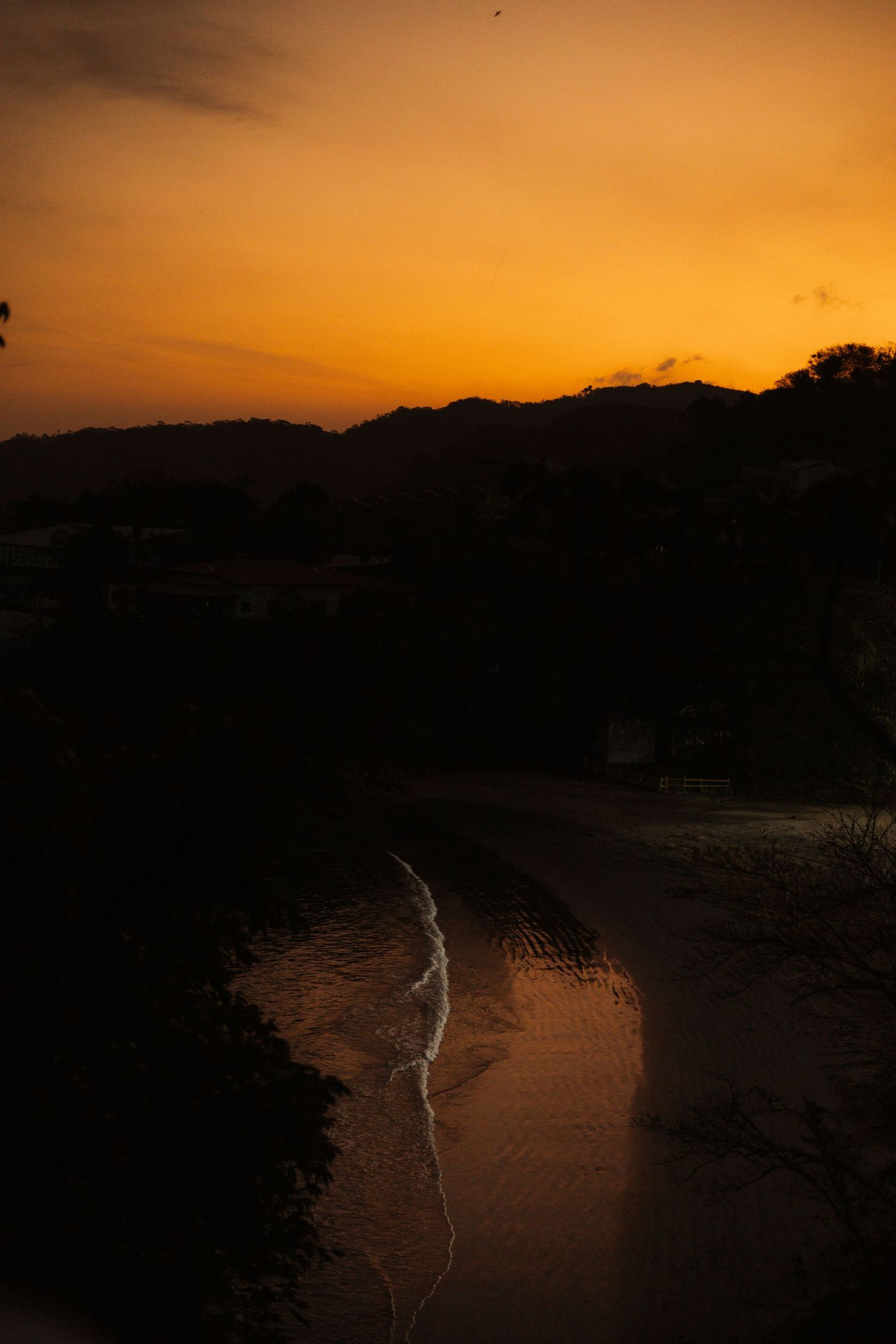 A dramatic orange sunset silhouettes mountains and coastline over a calm bay.