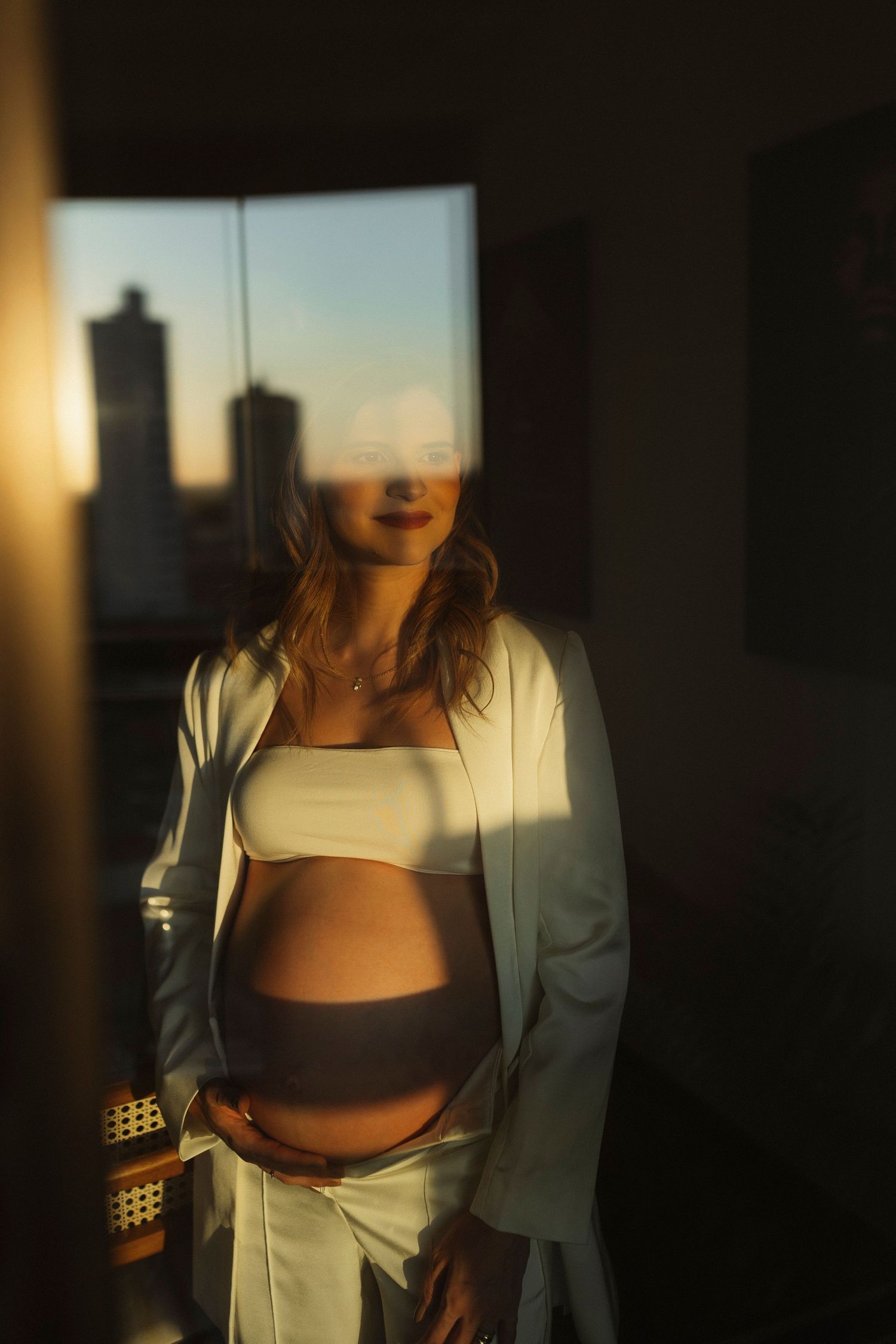 Maternity photoshoot in warm sunset light through window with dramatic shadows.