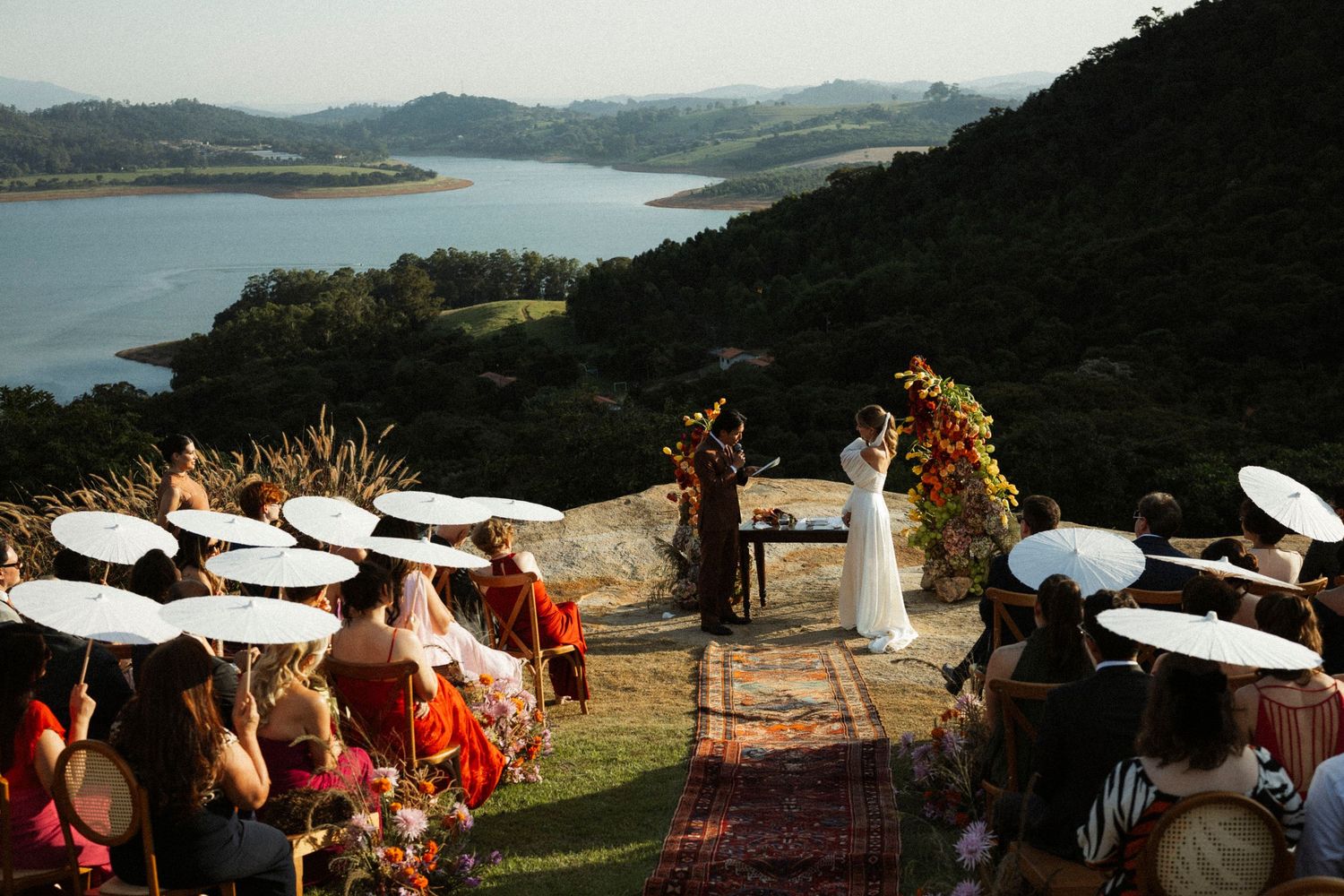 Outdoor wedding ceremony on a hillside overlooking a scenic lake and mountains, guests holding white parasols.