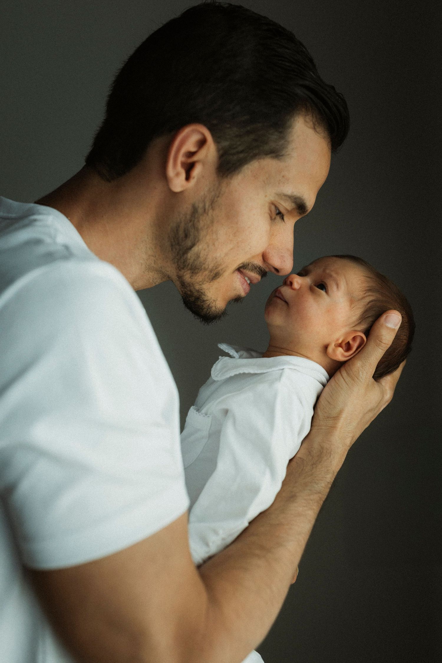 A father tenderly holds his newborn baby face-to-face, sharing an intimate bonding moment against a gray background.