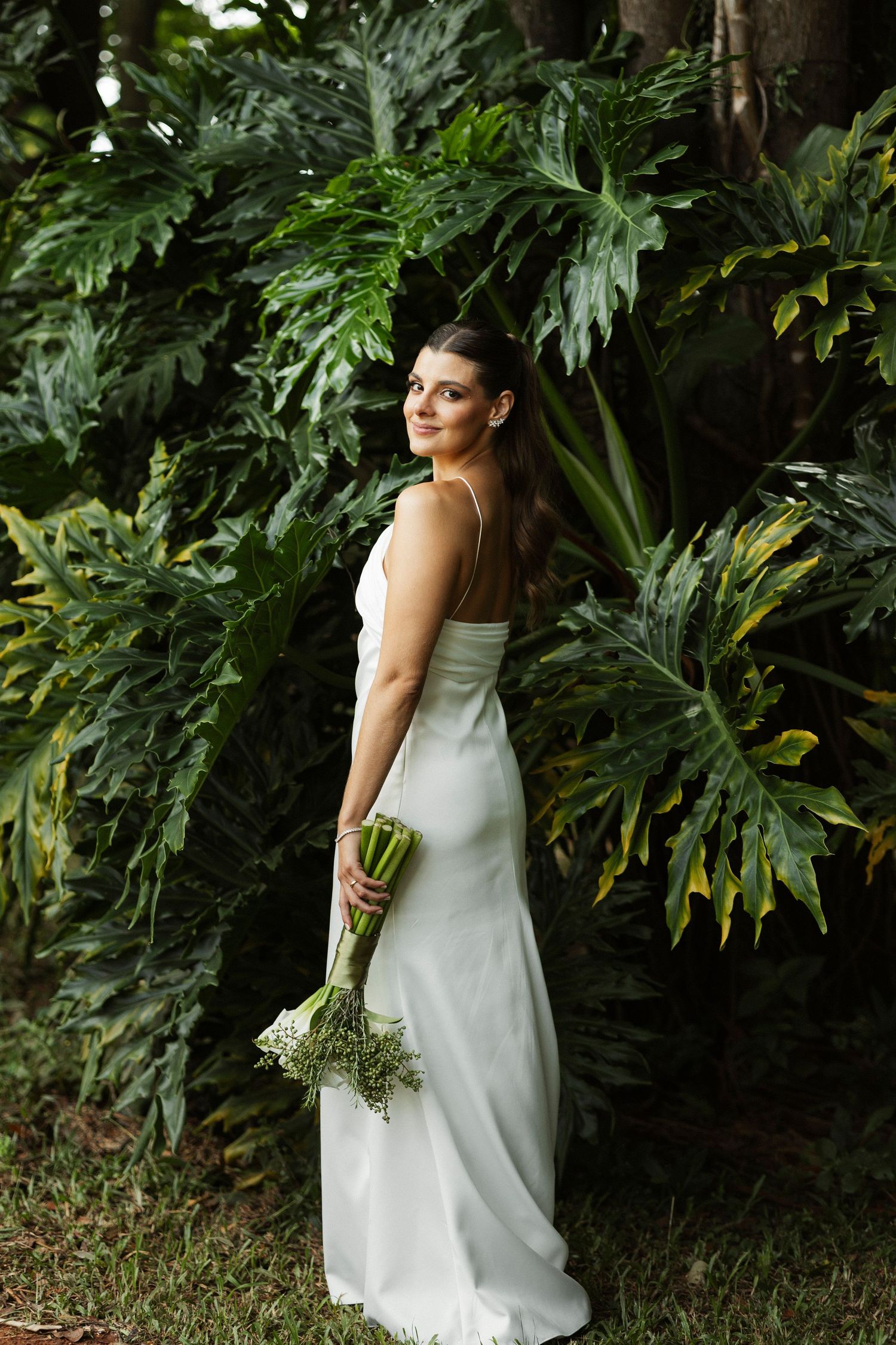 Bride in white satin gown holding green bouquet, posing among lush tropical foliage.