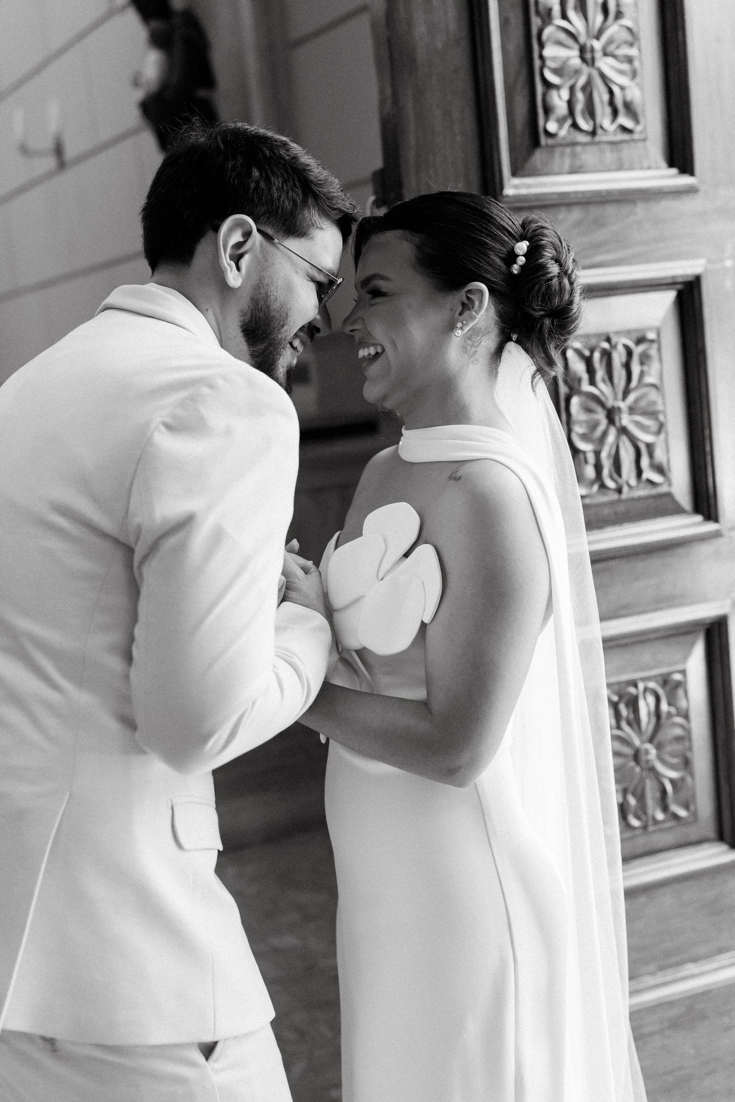Black and white photo of a laughing bride and groom in white attire sharing a joyful moment by an ornate door.