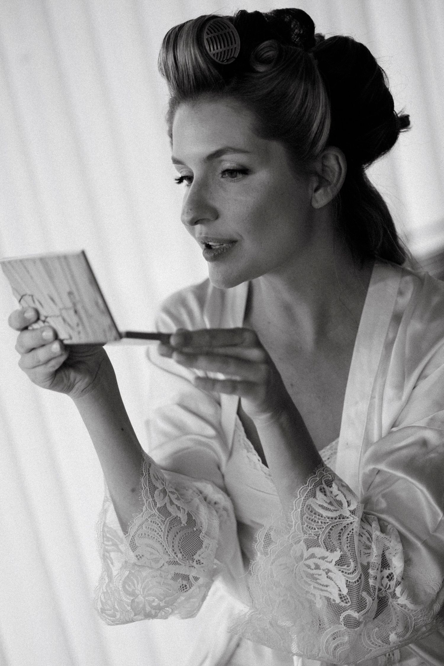 Bride in lace robe with hair rollers applying makeup with a brush while looking in a small mirror.