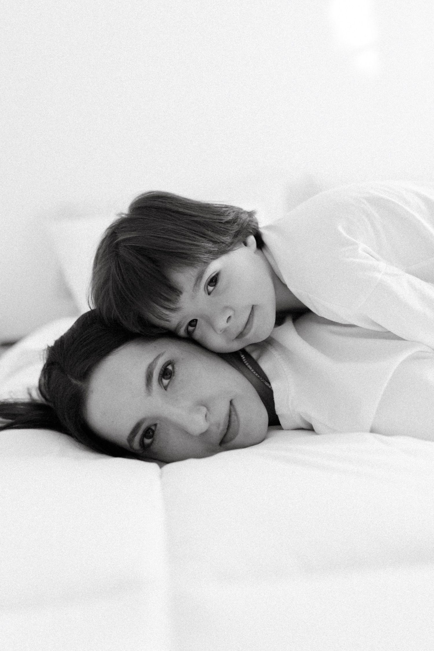 Black and white photo of a mother lying on a bed with her young child resting their head on her back.