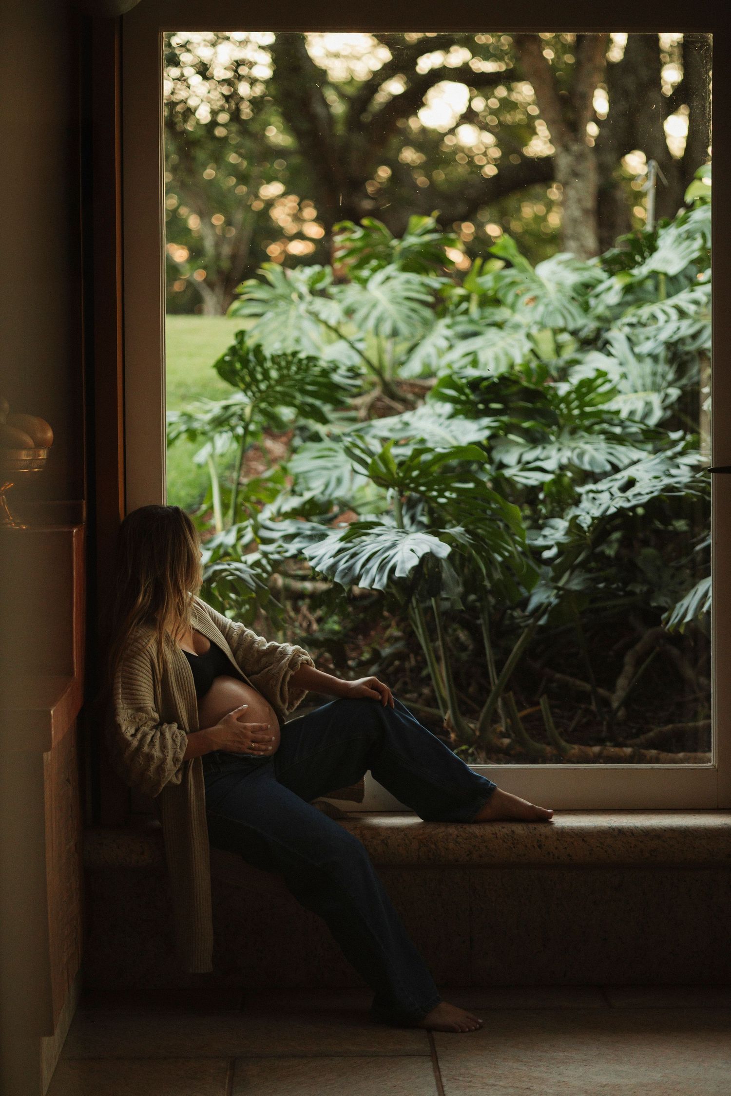 Silhouette of pregnant woman sitting by window with lush tropical plants outside.