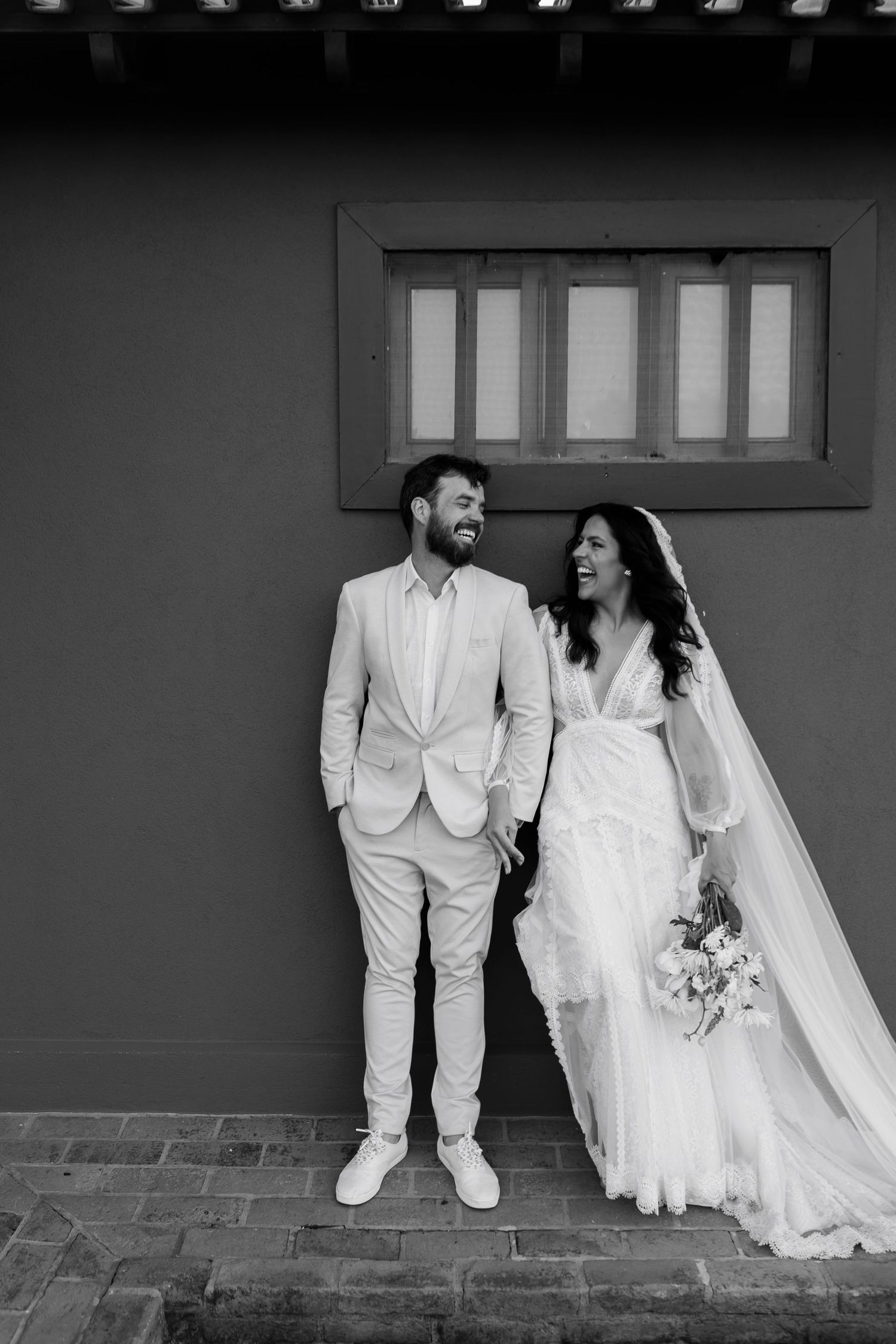 Wedding couple in white outfits poses against grey wall for artistic black and white portrait.
