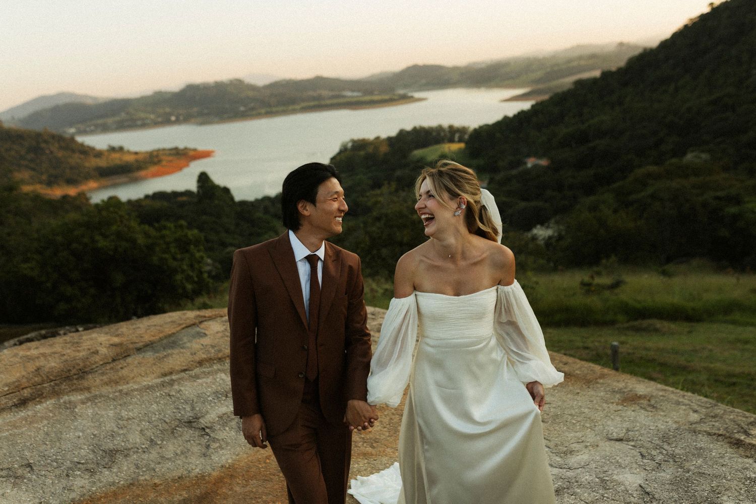 Couple in wedding attire holding hands on hilltop with scenic lake and green mountains at sunset.