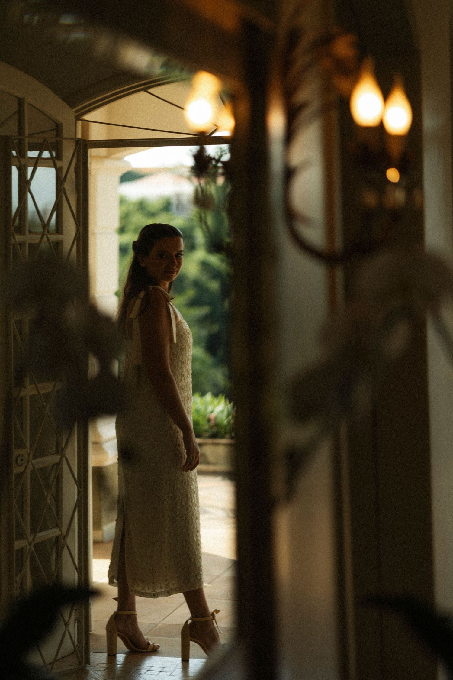 Woman in white lace midi dress and heels standing in a warmly lit doorway with garden views.