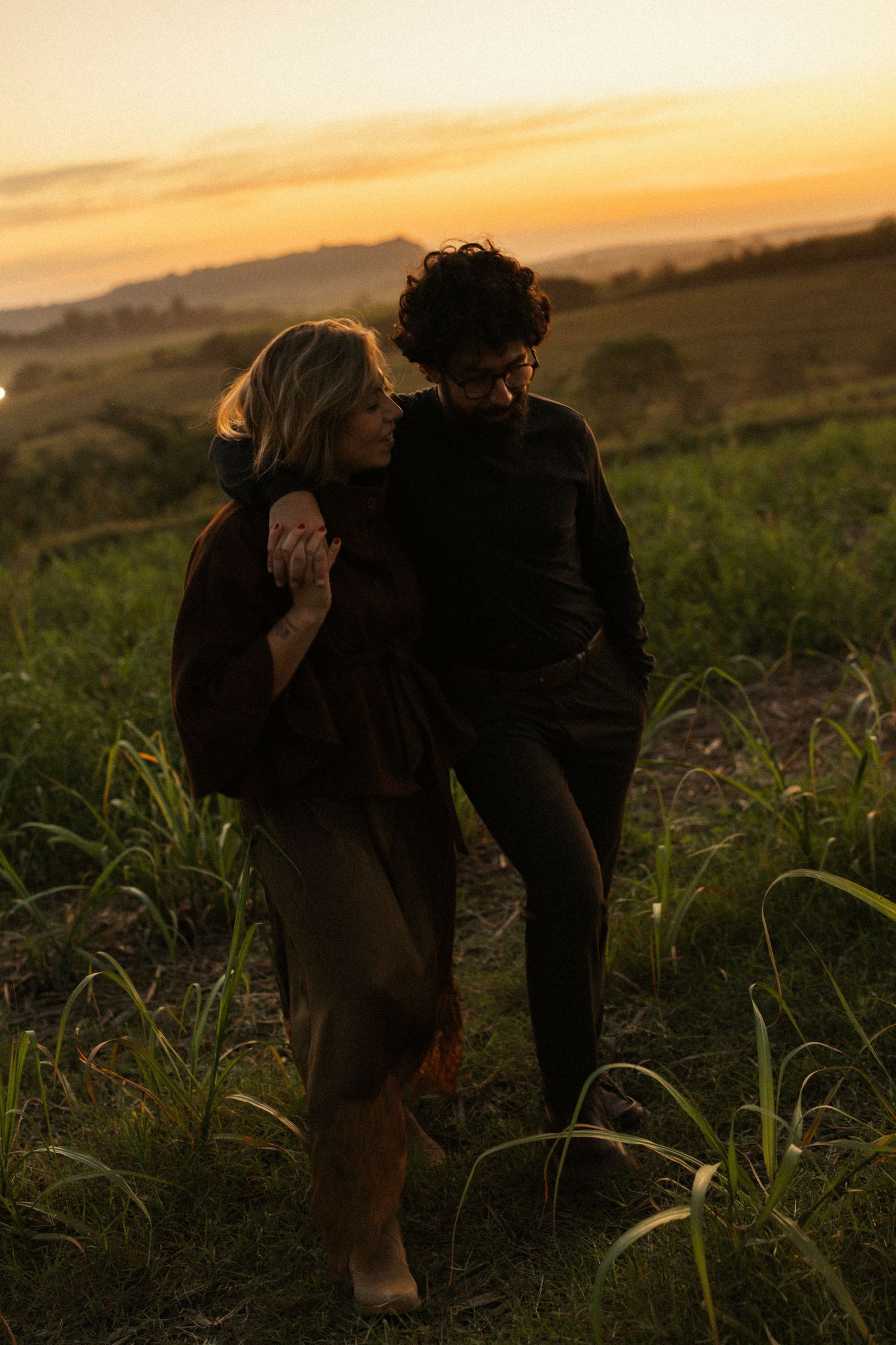 Couple embracing at golden hour sunset in lush green field, romantic outdoor portrait photography.